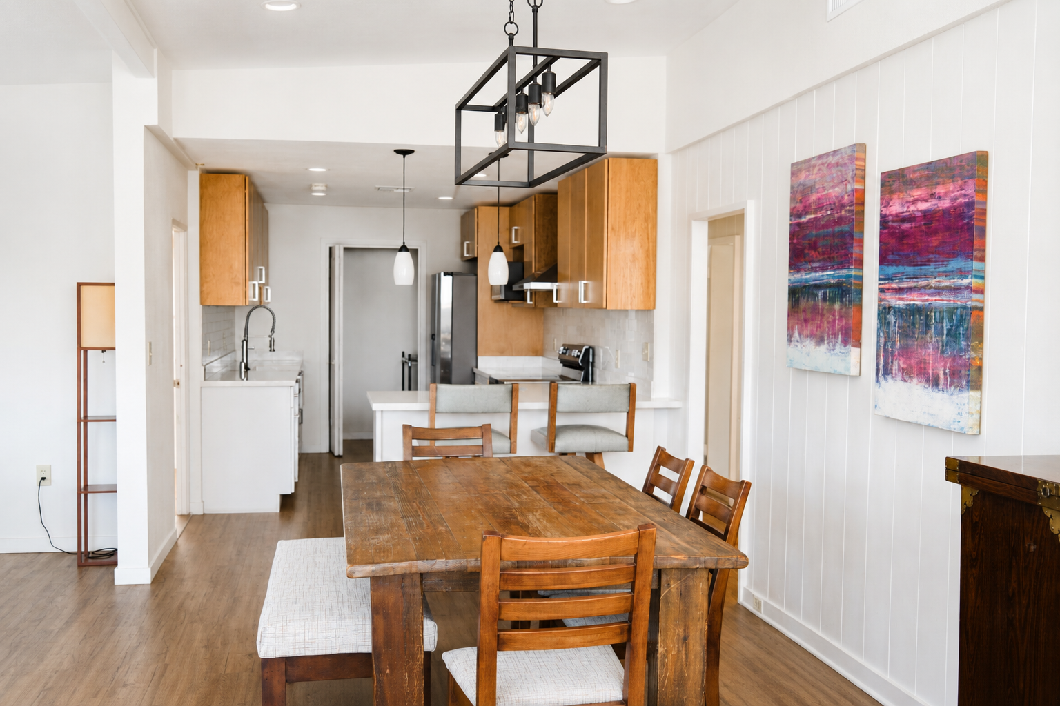A dining room and kitchen area with a wooden dining table and chairs, a white kitchen with wooden cabinets, artwork on white wall, and modern lighting fixtures.