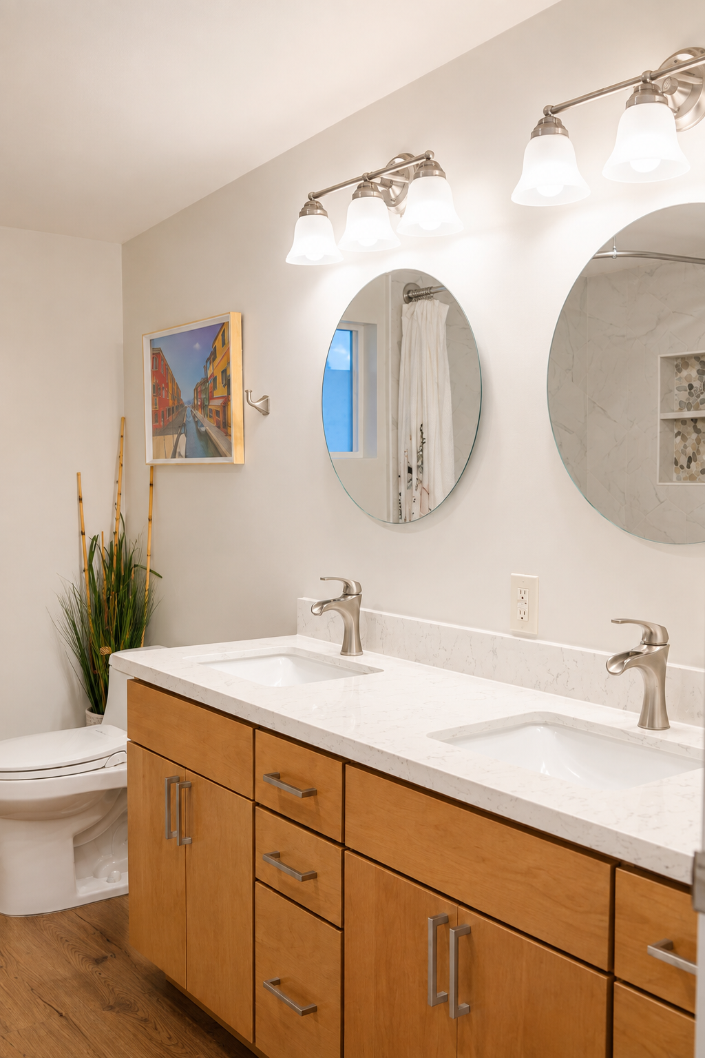 Modern bathroom with dual sinks, wooden cabinetry, oval mirrors, and a wall-mounted light fixture with four frosted glass shades. There is a small window, a toilet, and a plant with tall, green leaves and bamboo stalks in a white pot. A framed colorf