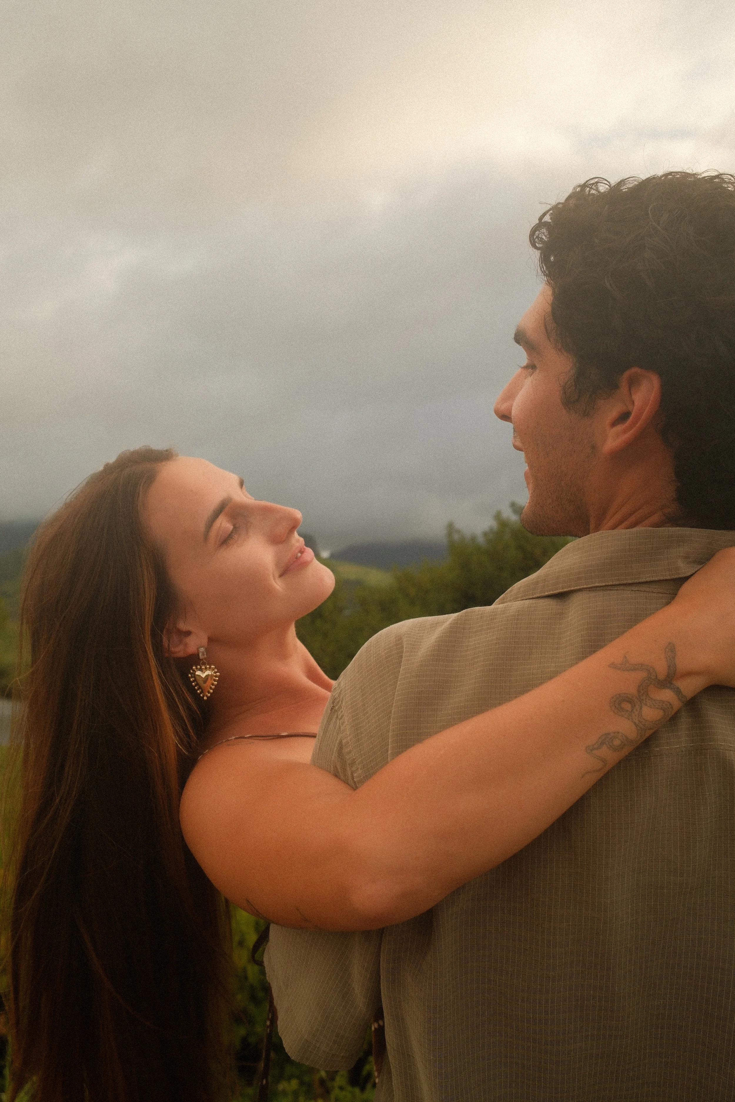 A couple dancing outdoors, looking into each other's eyes, with a cloudy sky and greenery in the background.