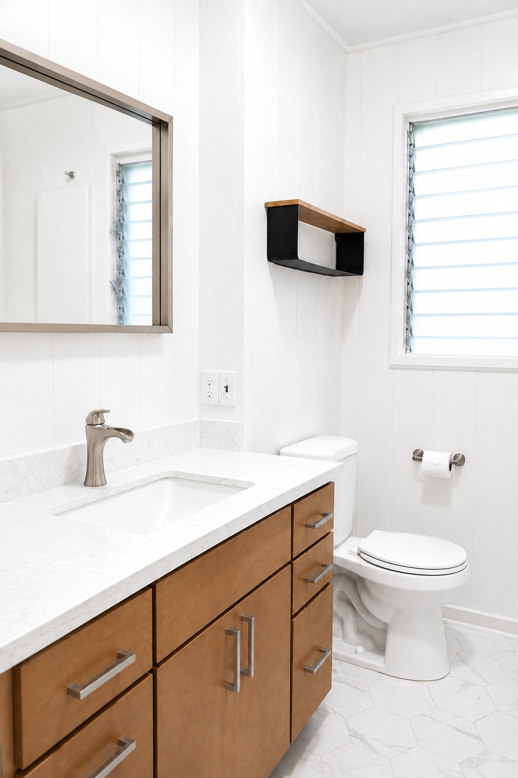 Bathroom with white walls, a wooden cabinet with silver handles, a white quartz countertop, a stainless steel faucet, a mirror with a silver frame, a small black and wood wall shelf, a toilet with a roll of toilet paper, and a window with louvers.