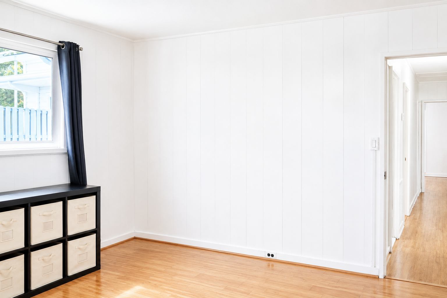 Empty room with white walls, wooden floor, a window with dark curtains, and a black storage unit with beige bins.