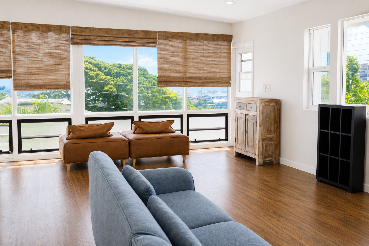 Living room with large windows, brown leather chairs, a gray sofa, a wooden cabinet, and a black shelf, overlooking a green outdoor landscape.