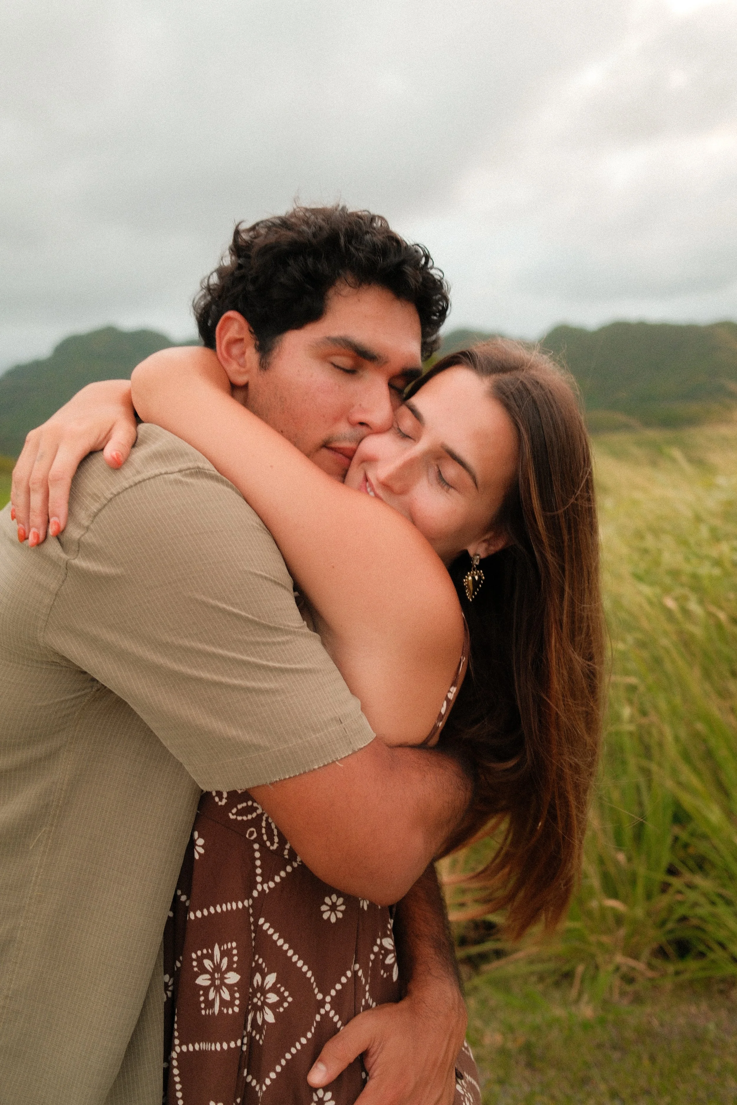A man and woman hugging outdoors in a grassy field with rolling hills in the background, both with their eyes closed and smiling.