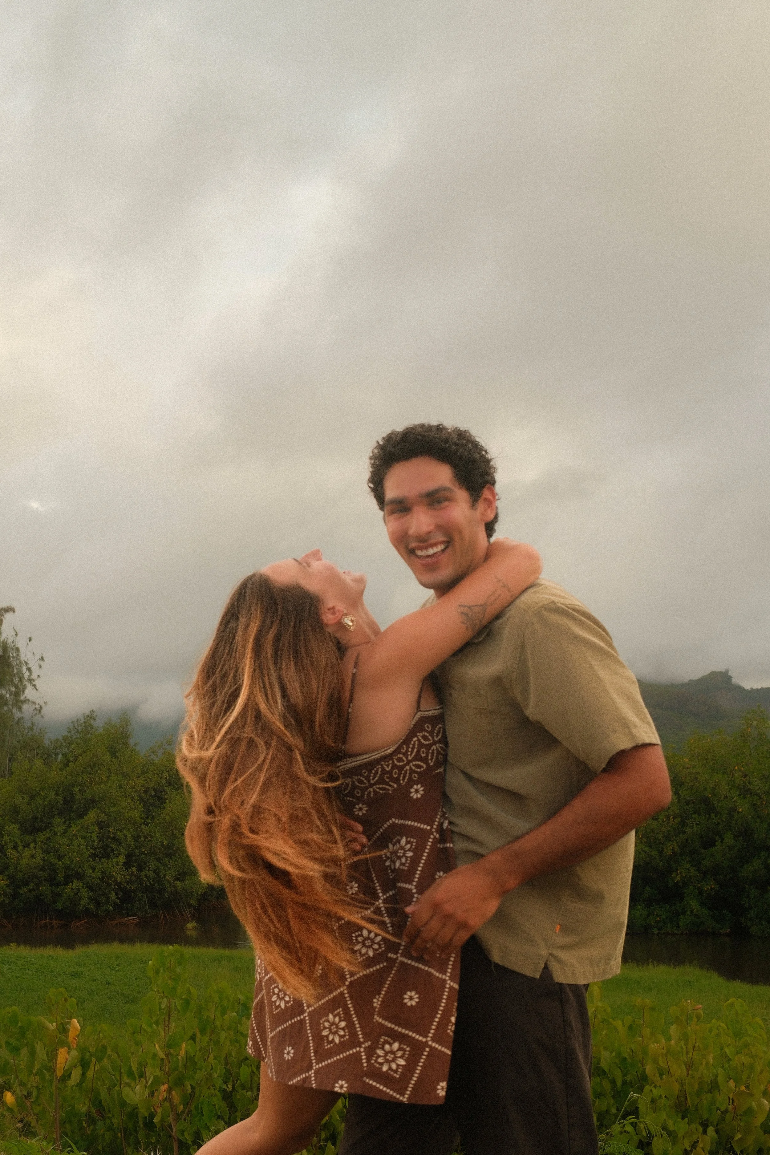 A young couple smiling and embracing outdoors near a river or lake, with lush greenery and mountains in the background on a cloudy day.