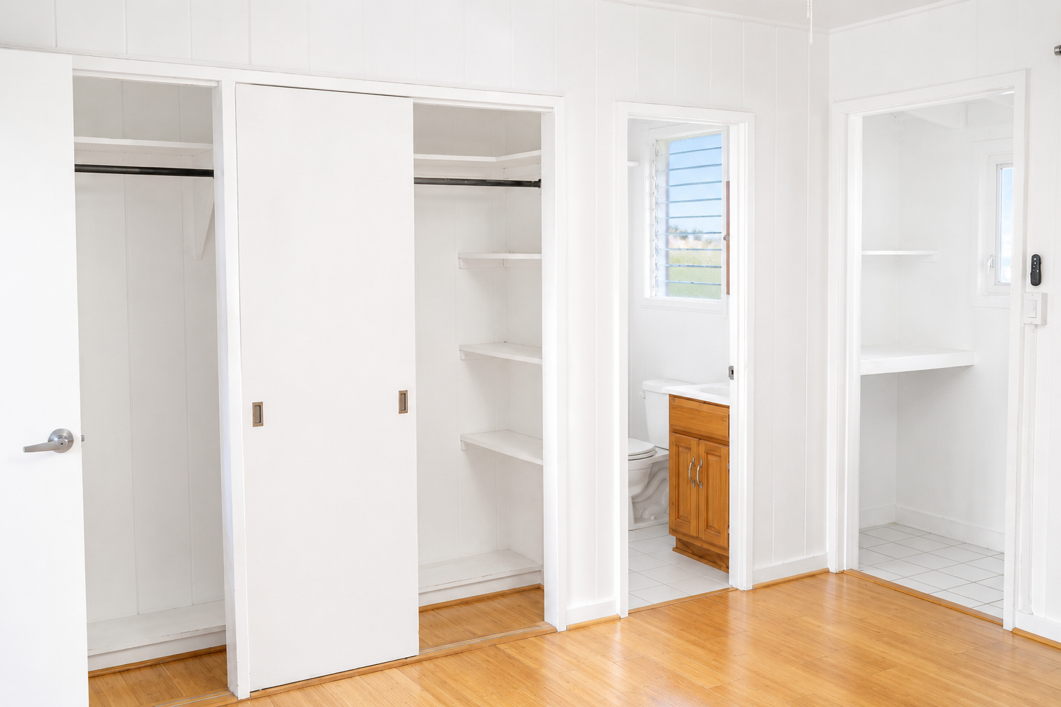 Empty closet with white sliding doors, a small bathroom with a wooden vanity and toilet, and a small alcove with a window, white walls and white tiled floors.