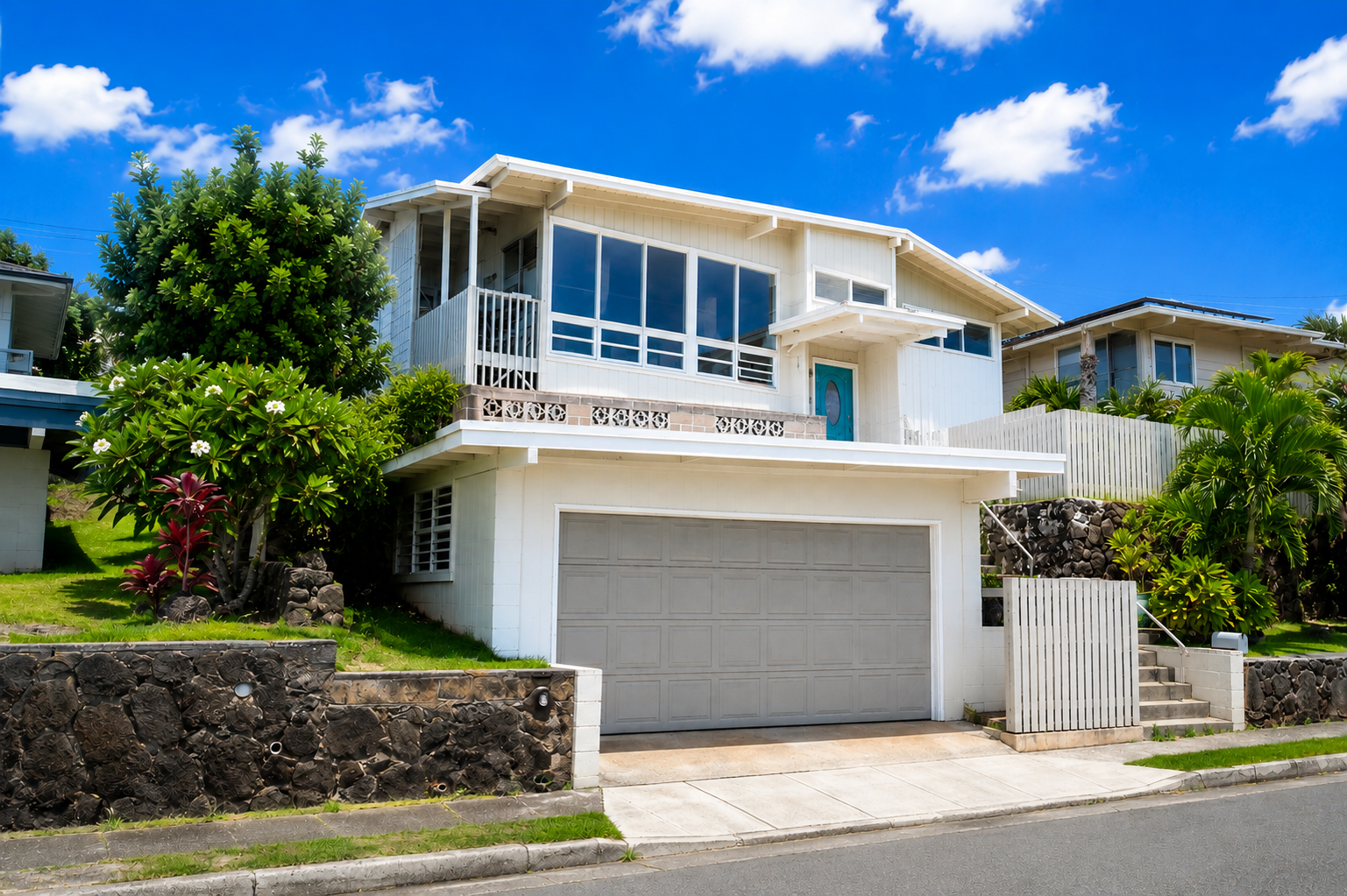 A white multi-story house with large windows, a garage, and surrounding greenery against a blue sky with clouds.