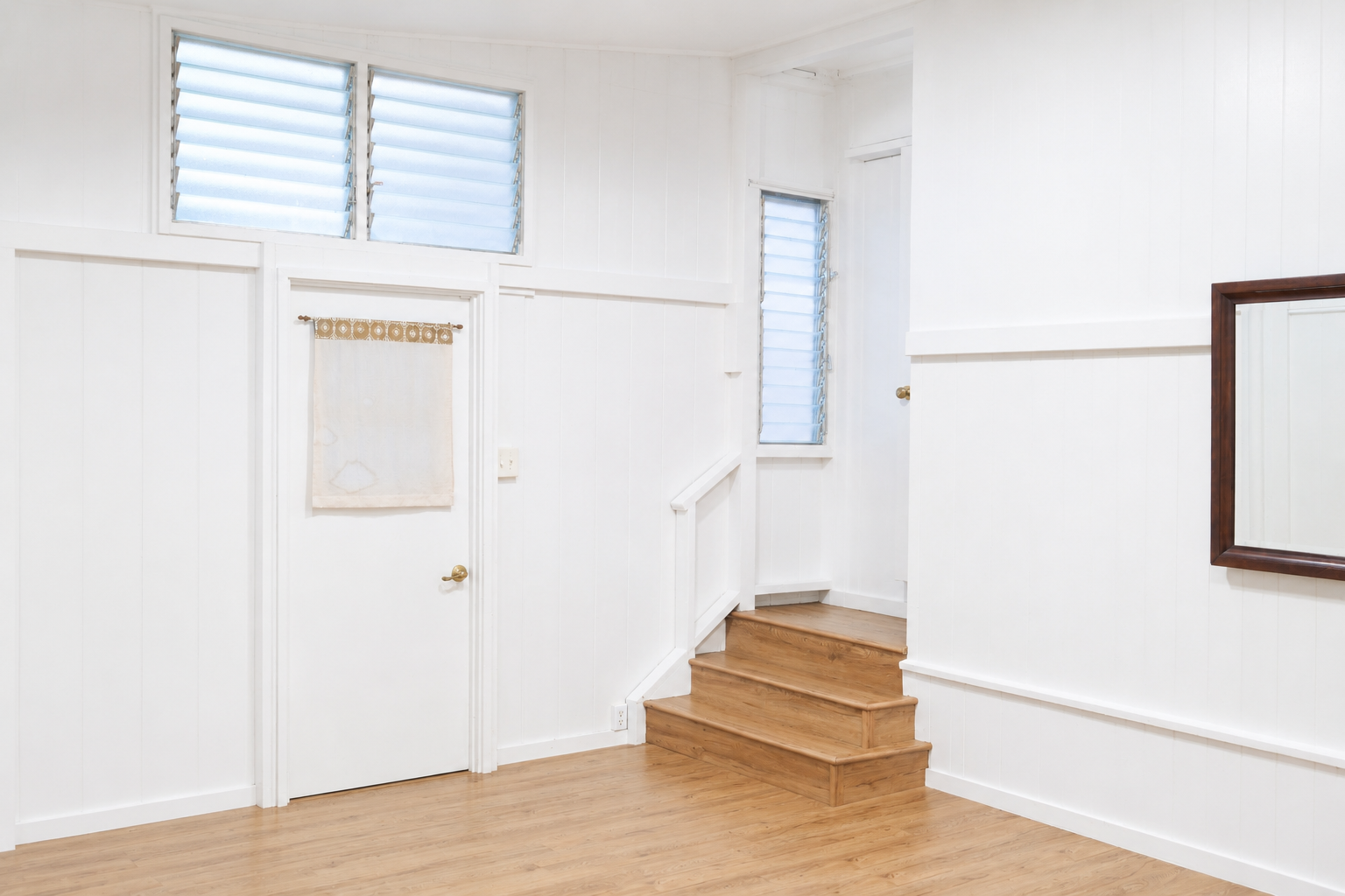 Interior of a bright room with white walls, wooden floors, a staircase with three steps, a small door with a curtain, two narrow windows with louvered blinds, and a framed mirror.