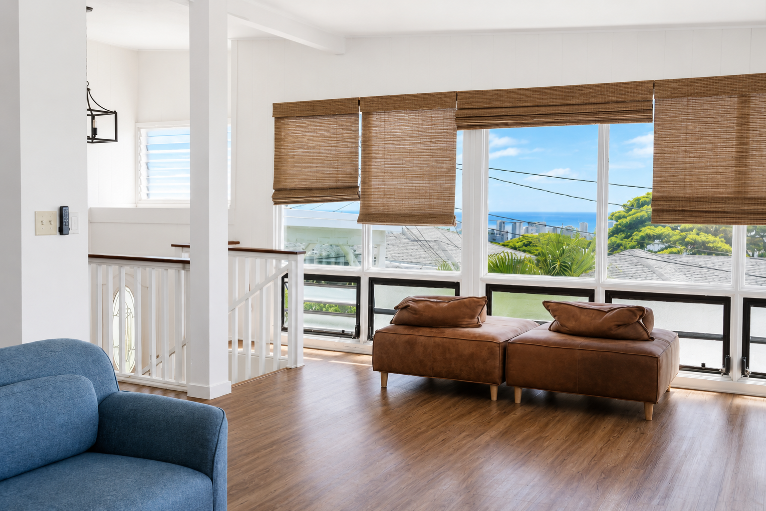 Living room with large windows showing ocean view, brown leather sofa, blue armchair, wooden flooring, bamboo roll-up window blinds, white walls, staircase railing.