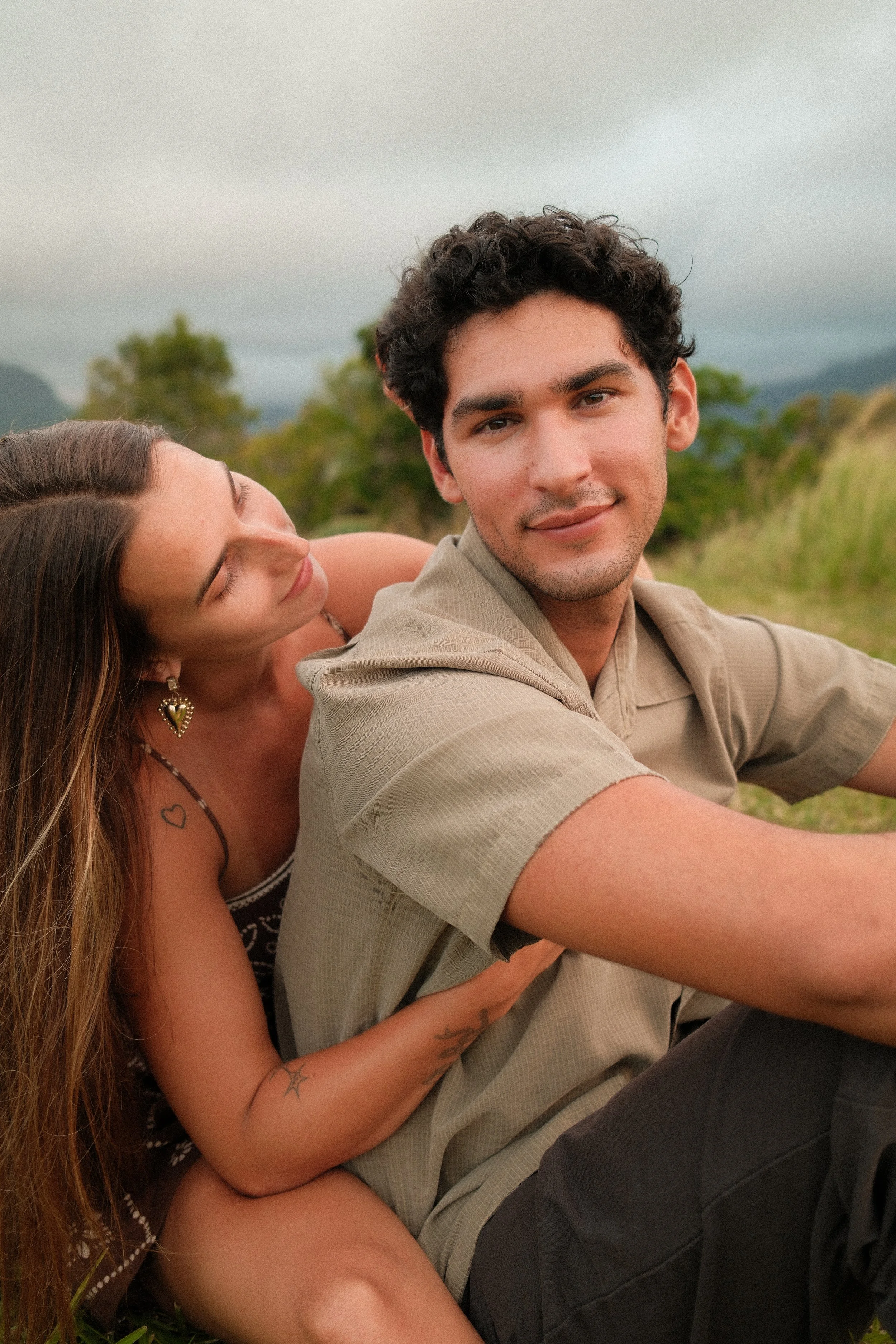 A young man with curly dark hair and light skin smiling while sitting outdoors in a grassy area with a woman with long brown hair and light skin leaning on his shoulder, both facing the camera. The background shows trees and cloudy sky.