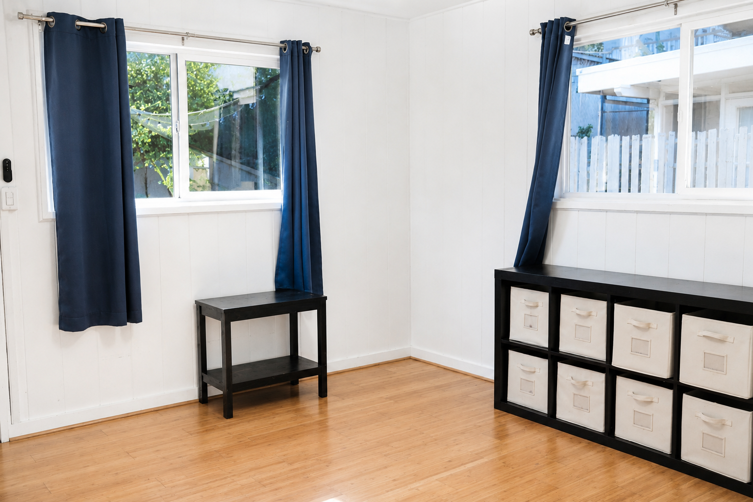 Empty room with white walls, hardwood floor, and two windows with blue curtains. Contains black furniture with white fabric storage bins.