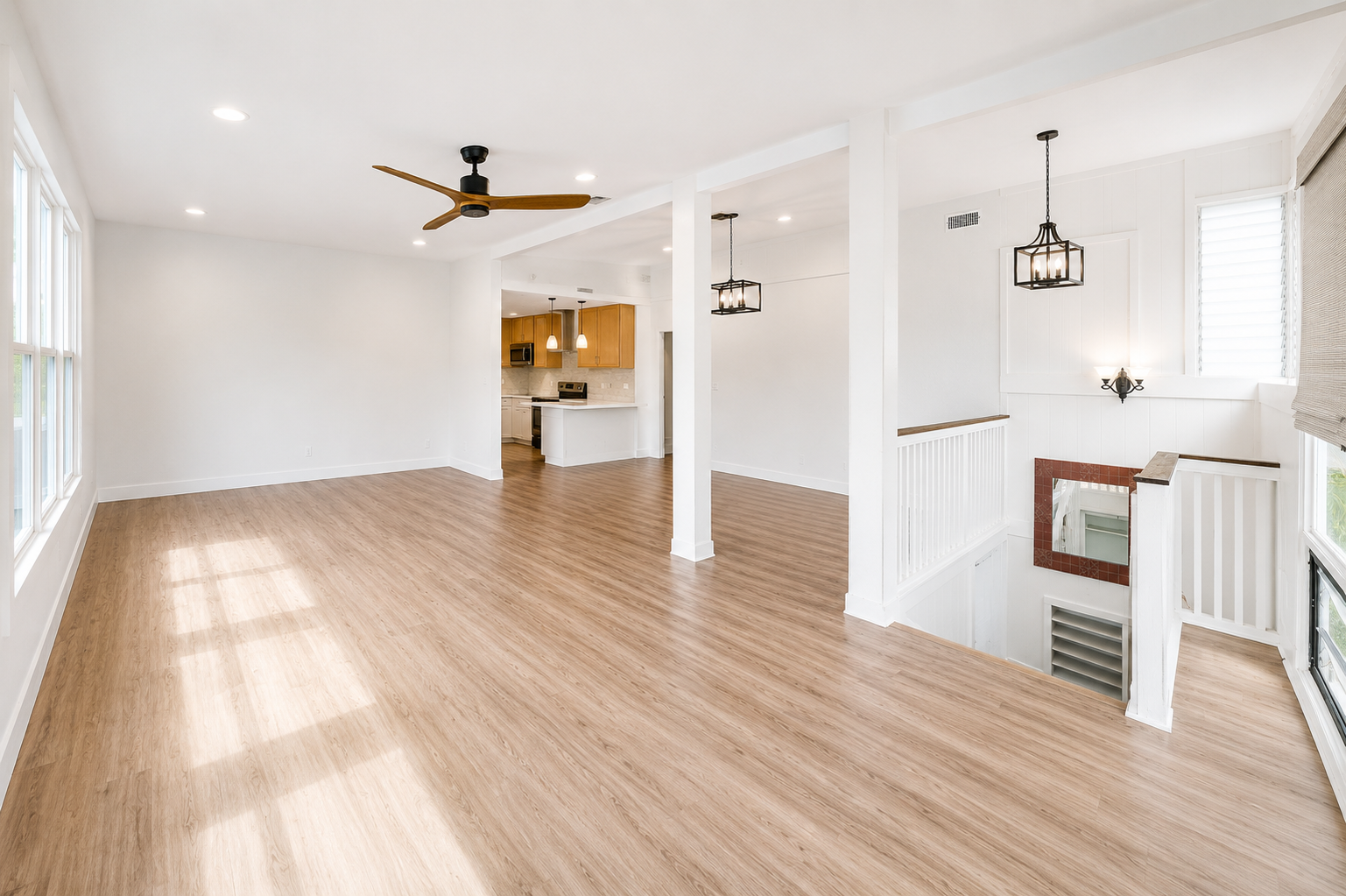 Hawaii Real Estate Photography: Spacious, empty living area with wood flooring, white walls, large windows, ceiling fan, and adjacent kitchen with wooden cabinets and island.