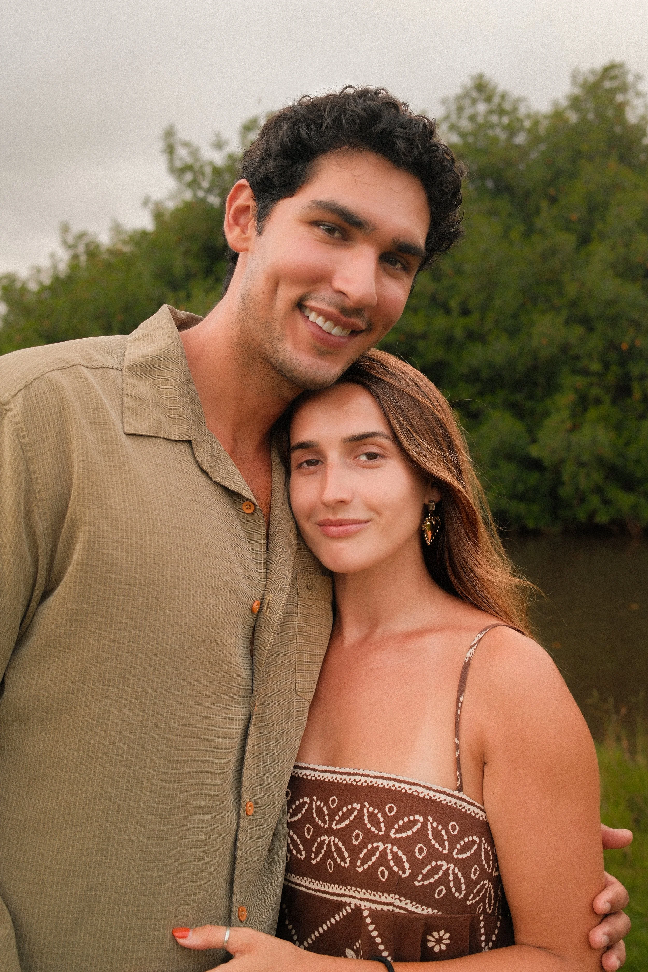 A young man with dark curly hair and a light beard smiling and hugging a young woman with long brown hair and earrings, standing outdoors with trees in the background.