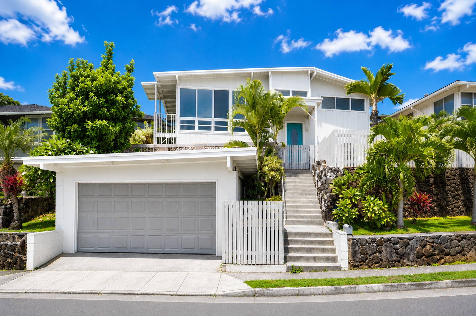 A modern white house with a garage, tropical plants, and stone landscaping under a blue sky with scattered clouds.