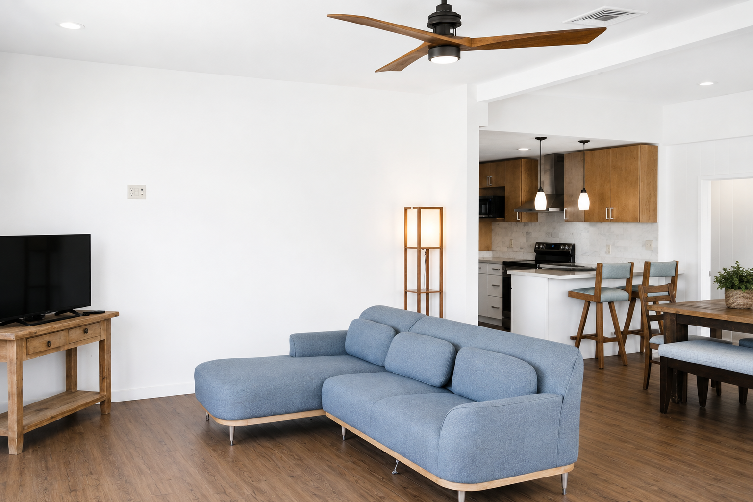 Living room with light blue sectional sofa, wooden TV stand with a flat-screen TV, and a white wall. In the background, there is a kitchen with wooden cabinets, a stove, and two hanging pendant lights. A wooden dining table with a potted plant is als