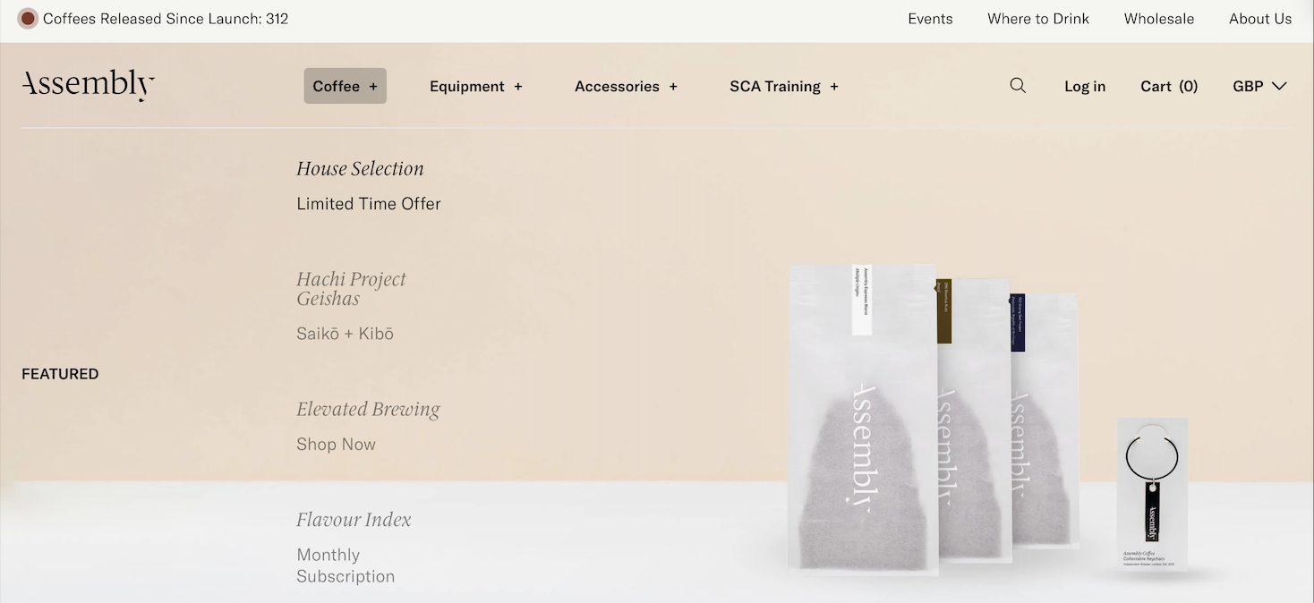 A website homepage for a coffee company named Assembly. The site features menu links at the top for events, where to drink, wholesale, and about us. There are options to log in, view cart, and select currency. The main content includes sections for house selection, the Hachi project Geishas, Elevated Brewing, and a flavor index. There are three white coffee bags and a black keychain with a white circular tag on the right side.