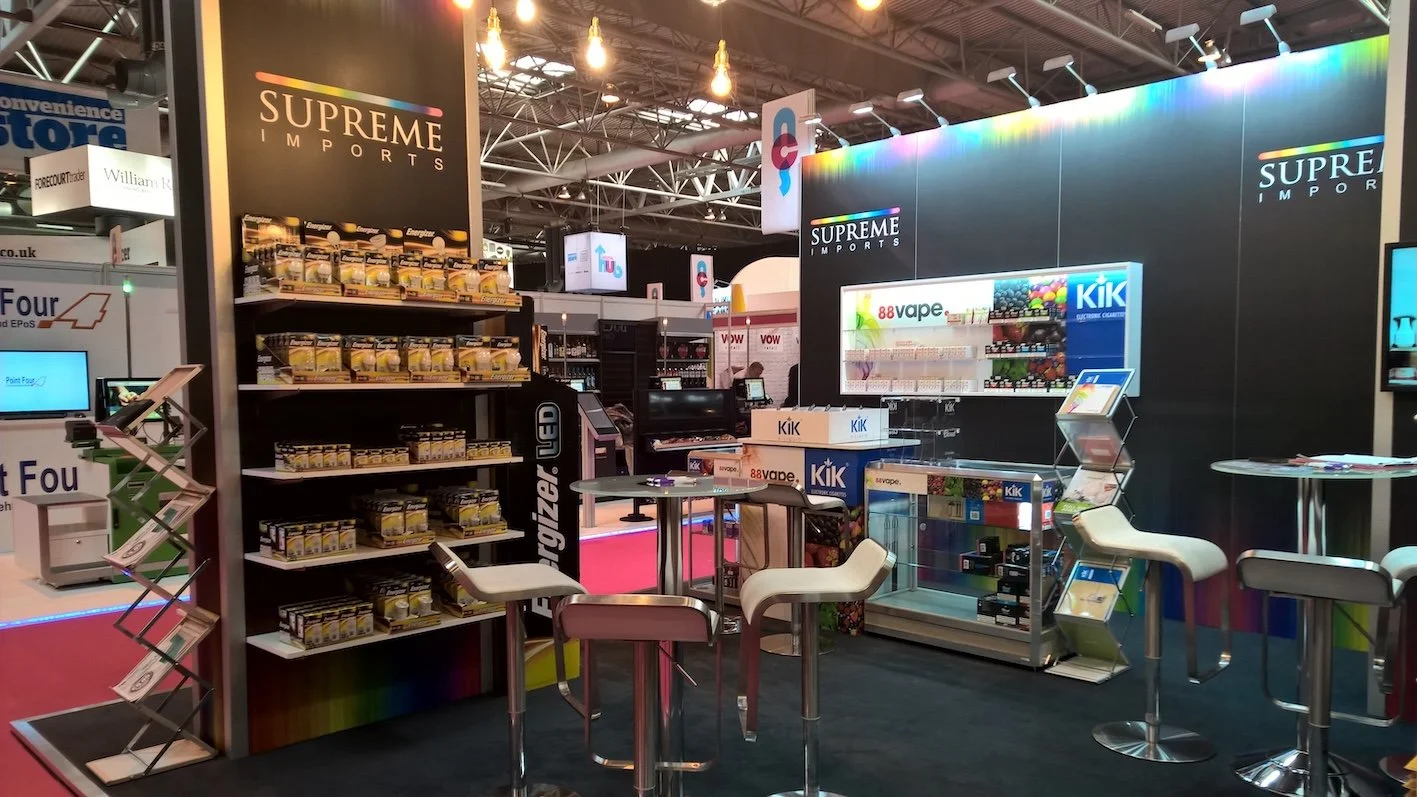Trade show booth for Supreme Imports featuring various vaping products, displays, and seating area with high chairs and tables.