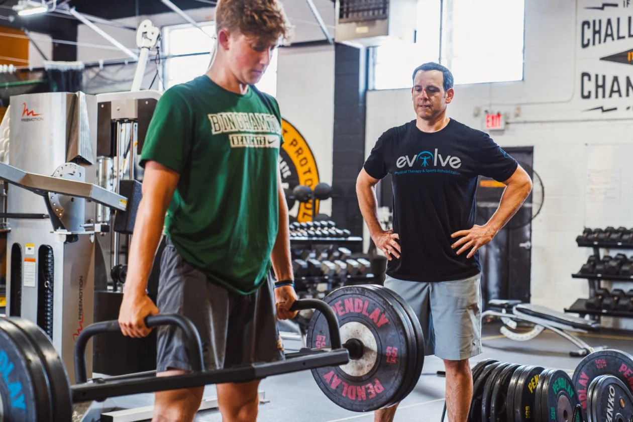 A young man is lifting a barbell with weights during a workout in a gym, while a trainer or coach observes him and provides guidance.