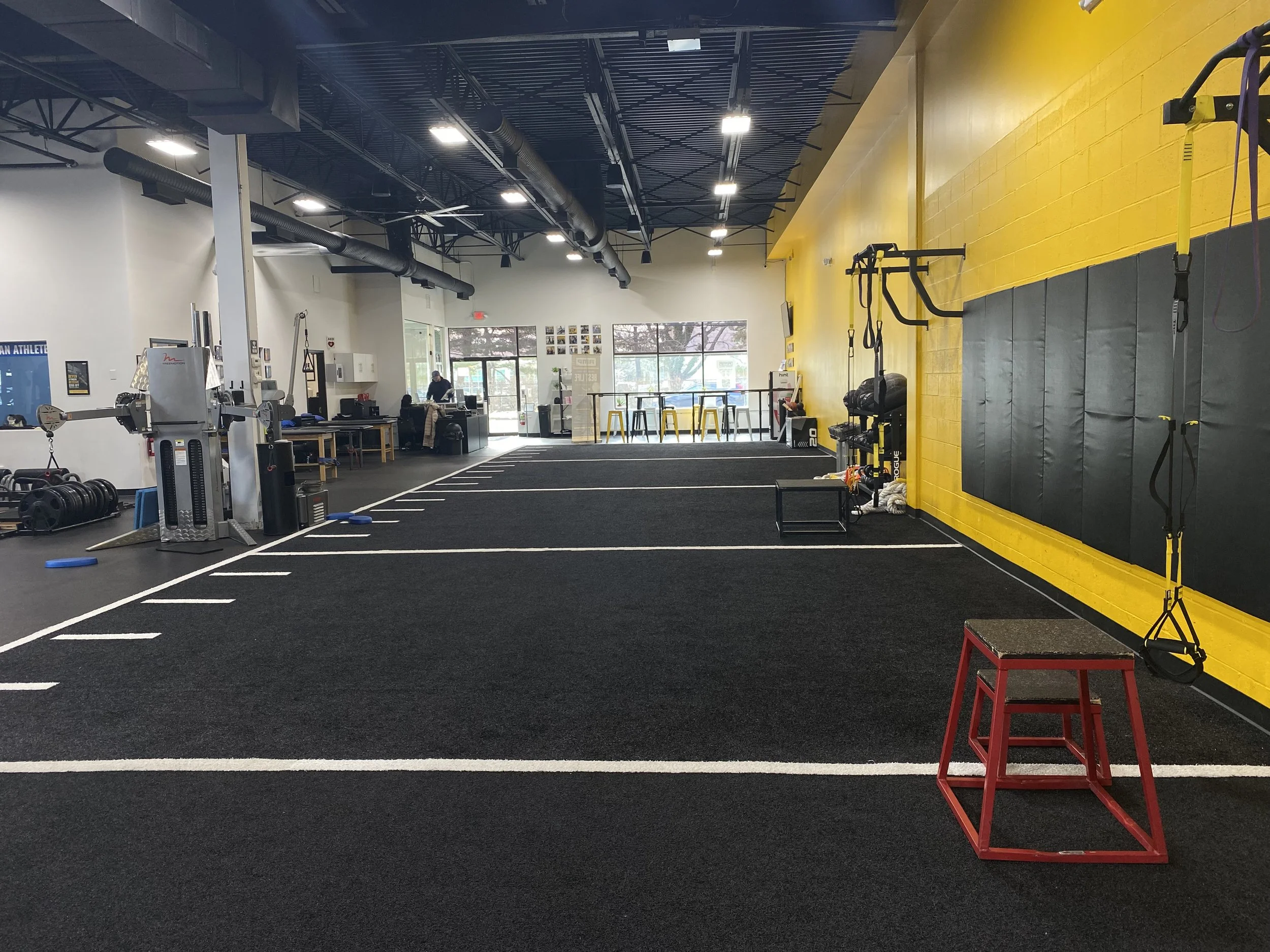 Interior view of a modern gym with black rubber flooring, white walls, and a yellow accent wall. Equipment includes weights, a sled, and resistance bands. There are weightlifting stations, a small seating area with yellow stools, and a few people wor