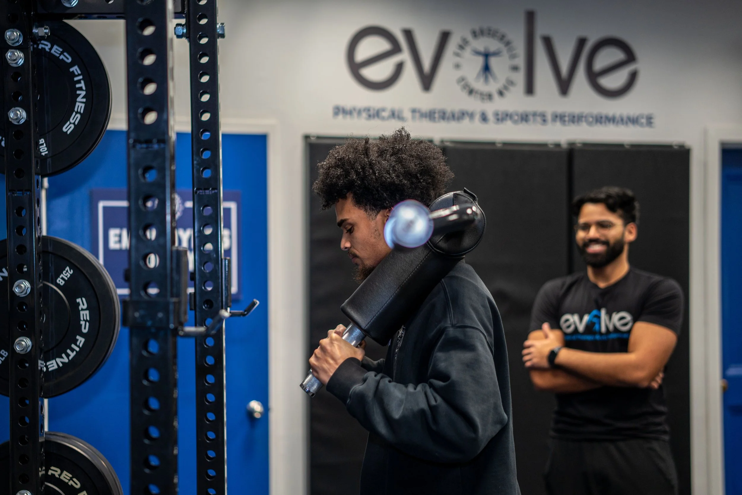 A man lifting a large weight on his shoulder in a gym, while a smiling trainer stands in the background with arms crossed, inside a gym labeled Evolve Physical Therapy & Sports Performance.