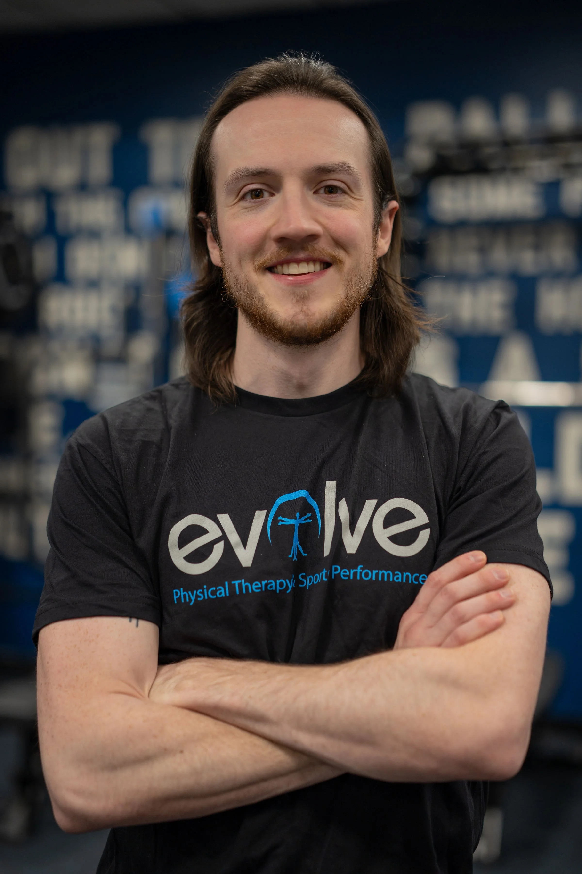 A man with long brown hair, beard, and light skin, smiling with arms crossed, wearing a black T-shirt with blue text and graphics, in a gym or fitness center with blue walls and shelves of equipment in the background.