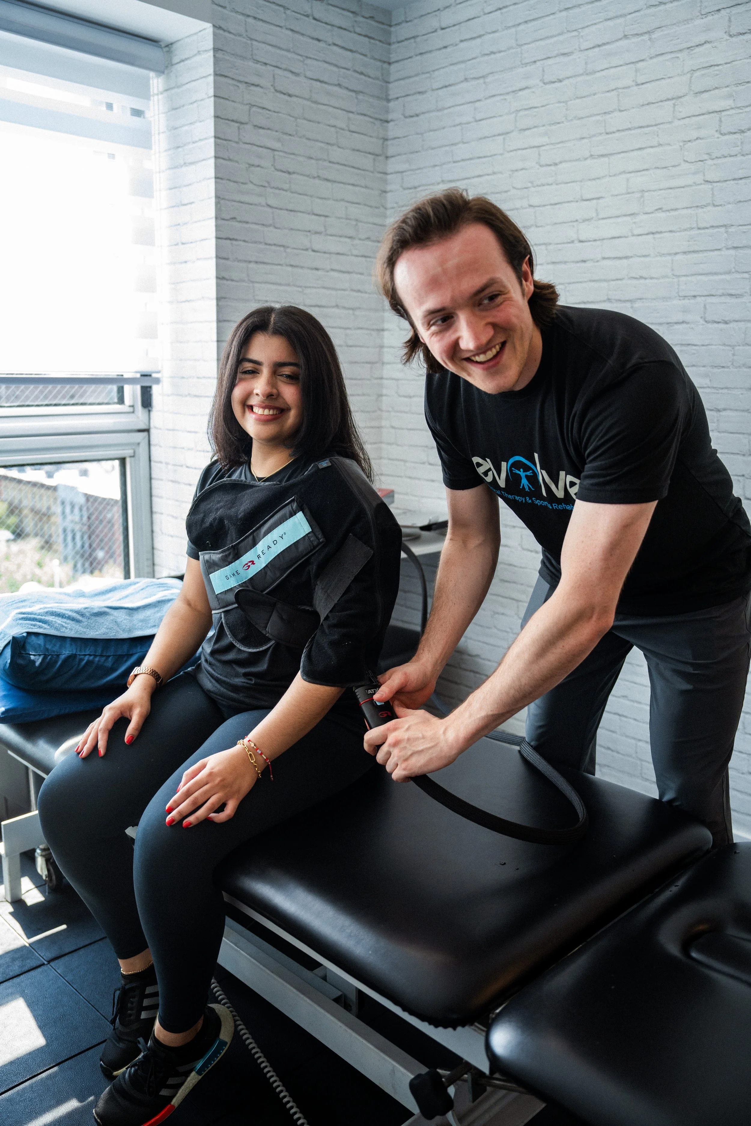 A physiotherapist adjusting a therapy band on a smiling woman sitting on a treatment table in a clinic with white brick walls and a window.