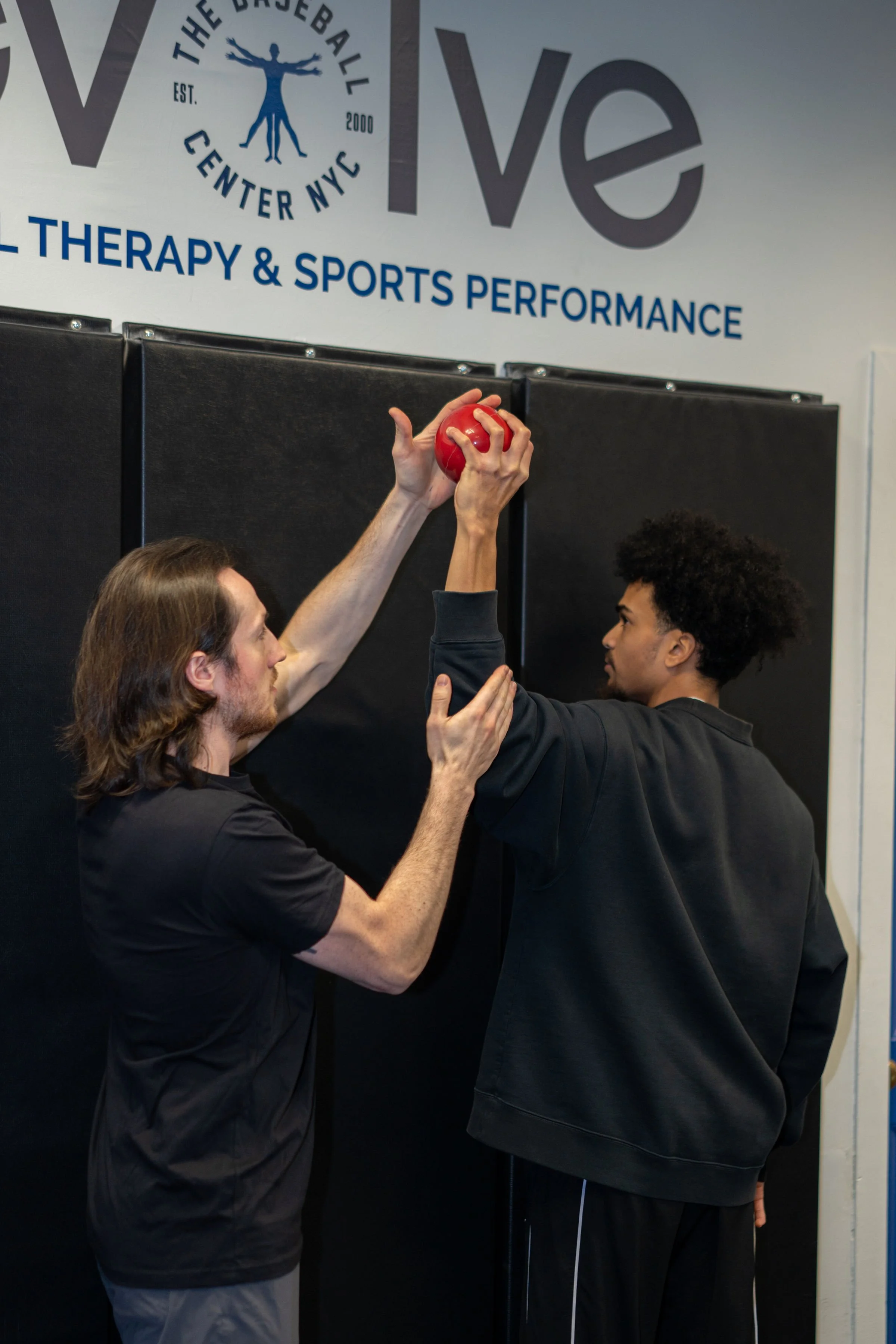 Two men are practicing a shoulder pass with a red medicine ball in a gym, with a black padded wall and a sign that reads 'The Baseball Center NYC' and 'Evolve Physical Therapy & Sports Performance' in the background.