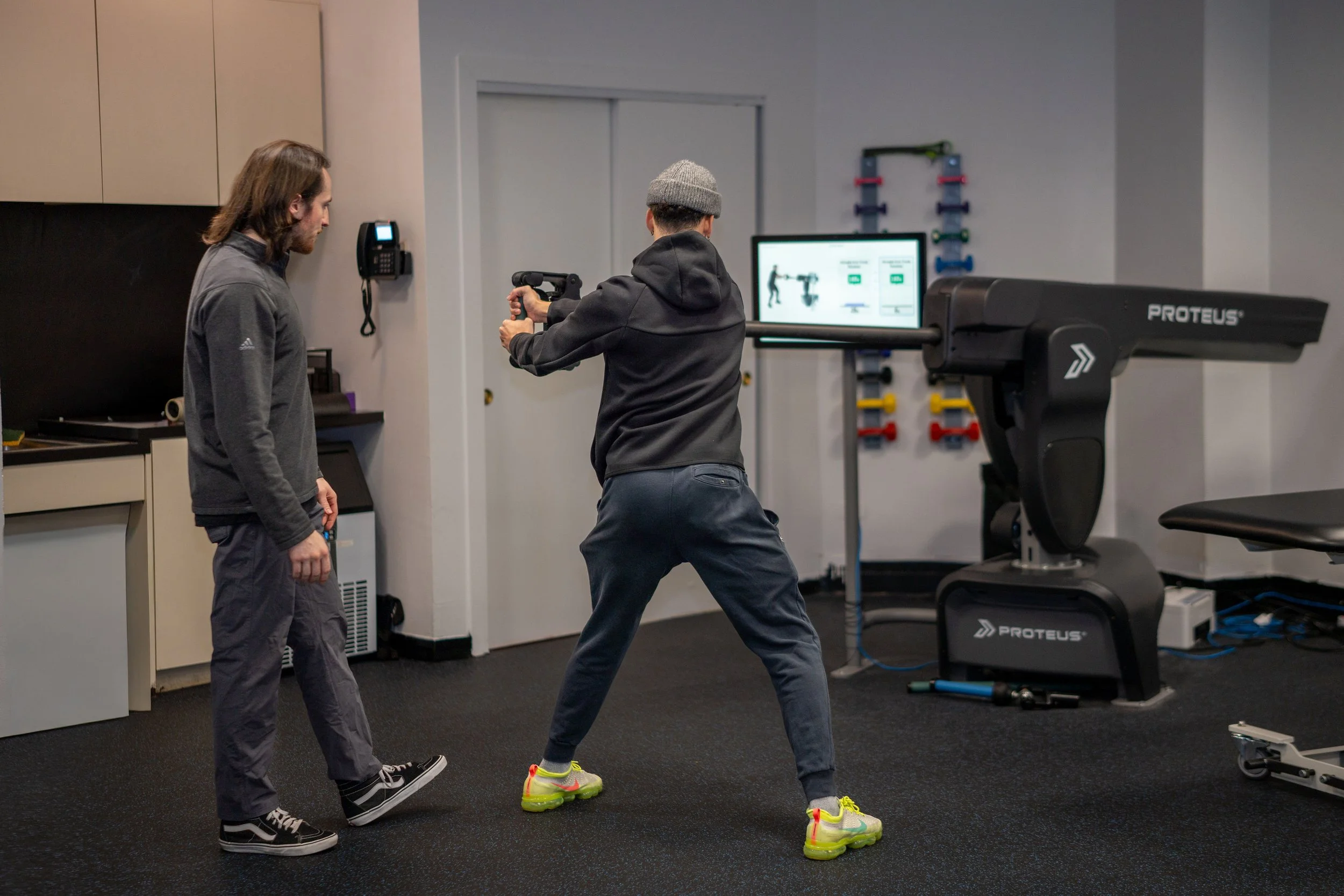A person in athletic attire and colorful sneakers performing an exercise with a gun-shaped device, monitored by another individual, in a gym setting with exercise equipment and a computer screen in the background.