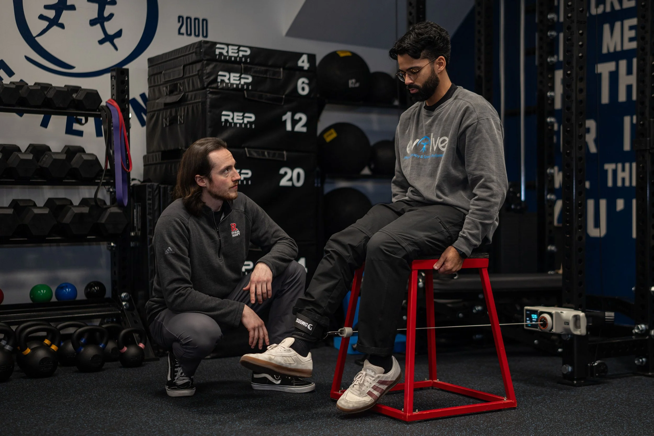 Two men in athletic wear in a gym, one sitting on a stool and the other kneeling on the floor, having a conversation. Gym equipment such as kettlebells, medicine balls, and weight plates are visible in the background.