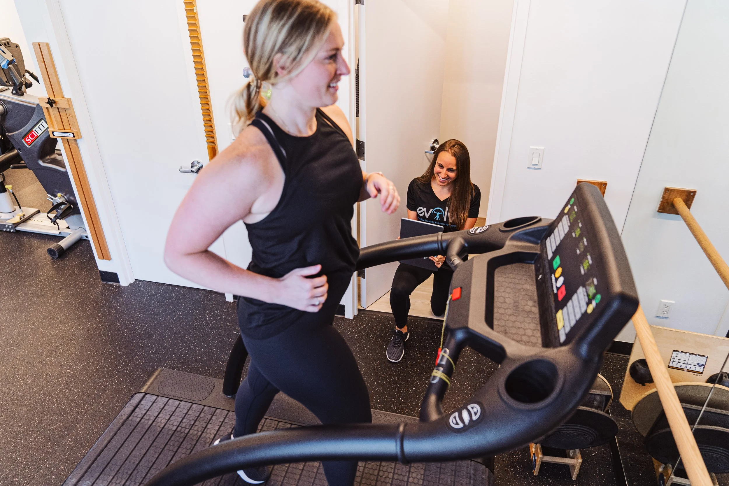 Two women in a gym, one running on a treadmill and the other sitting on the floor, smiling and holding a notebook, with exercise equipment visible in the background.