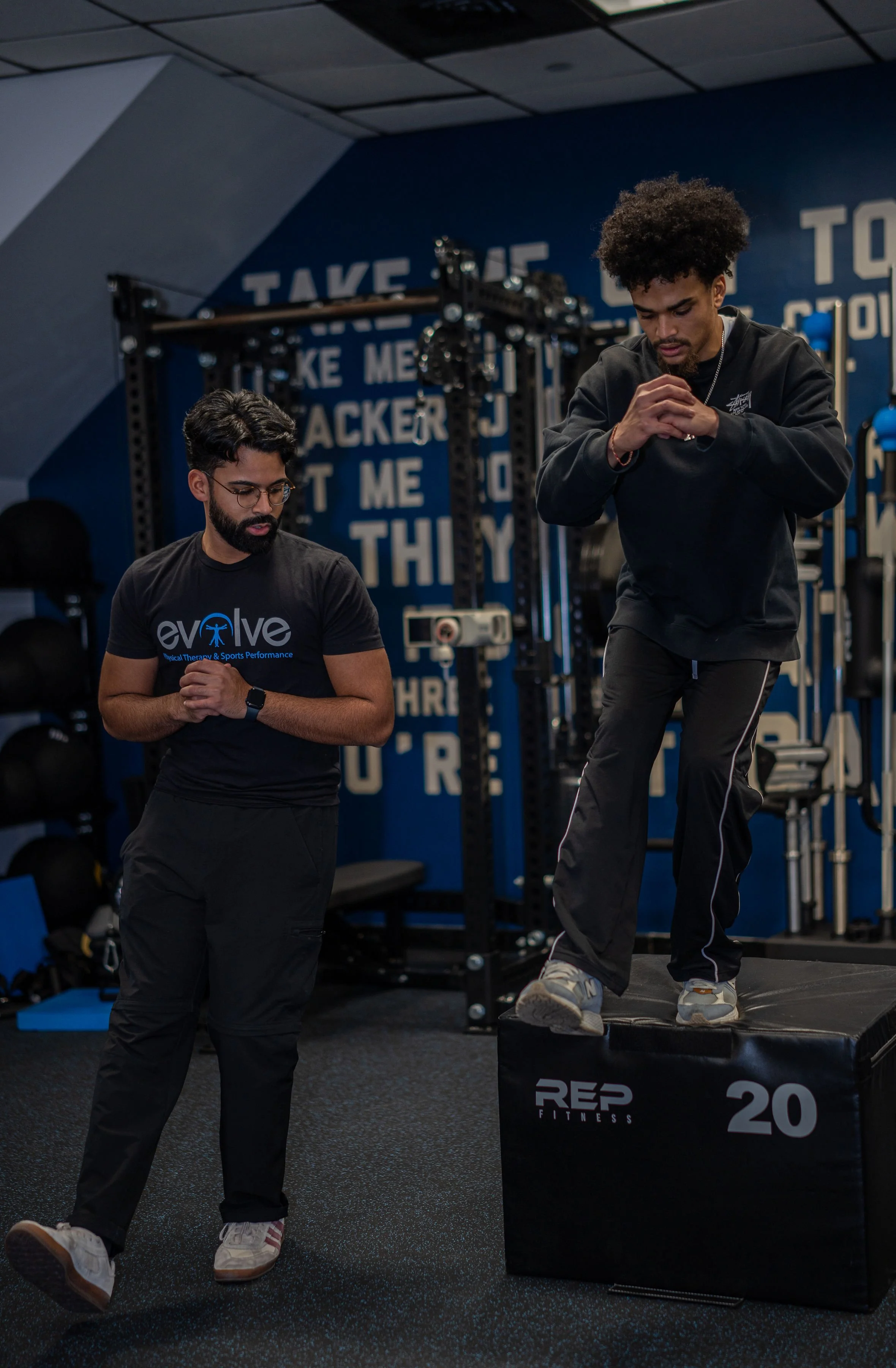A trainer watches a man jump onto a plyometric box while another trainer observes in a gym with motivational quotes on the wall.