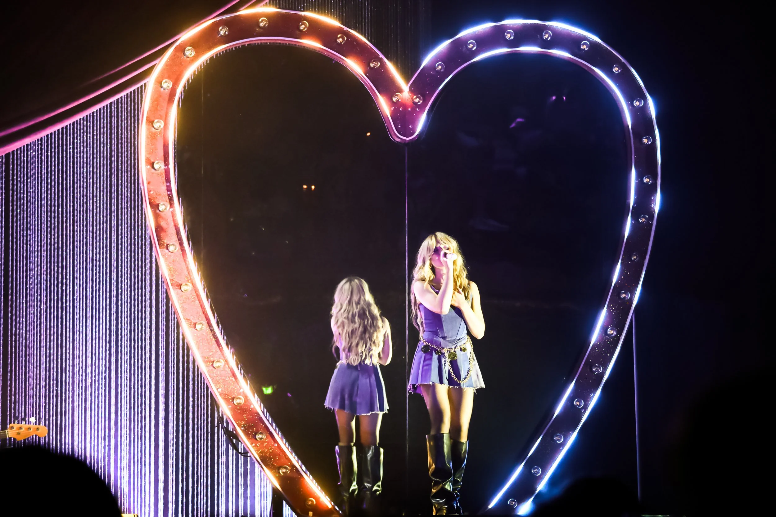 A female singer performs on stage in front of a large, illuminated heart-shaped mirror with bright edge lights. She has long hair, wears a sleeveless dress with a chain belt and knee-high boots. There is a young girl with long hair, also in a dress and boots, standing behind her reflected in the mirror.
