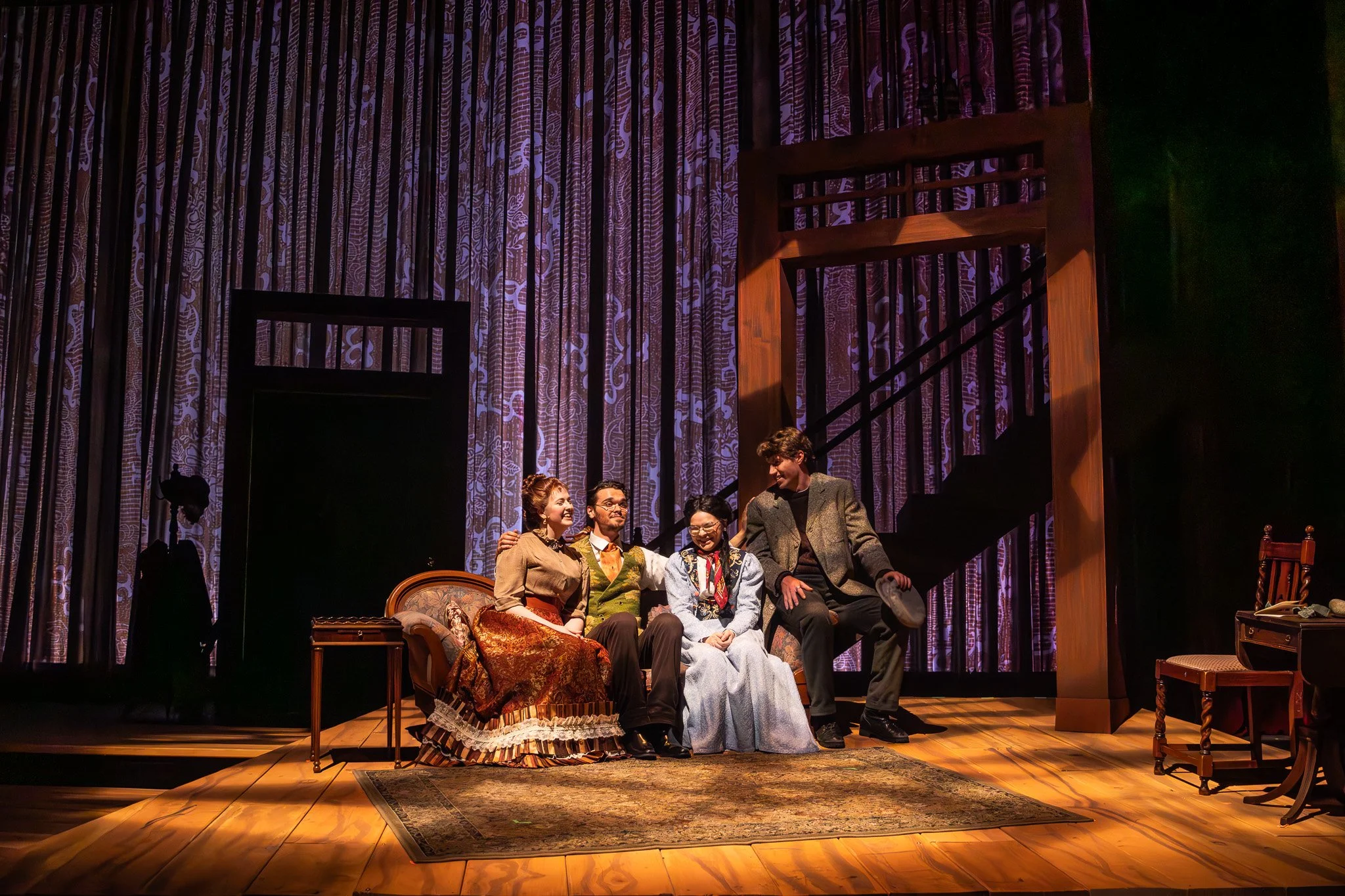 Theatre stage with actors in period costumes sitting on a sofa, set against a backdrop of a staircase and patterned curtains.