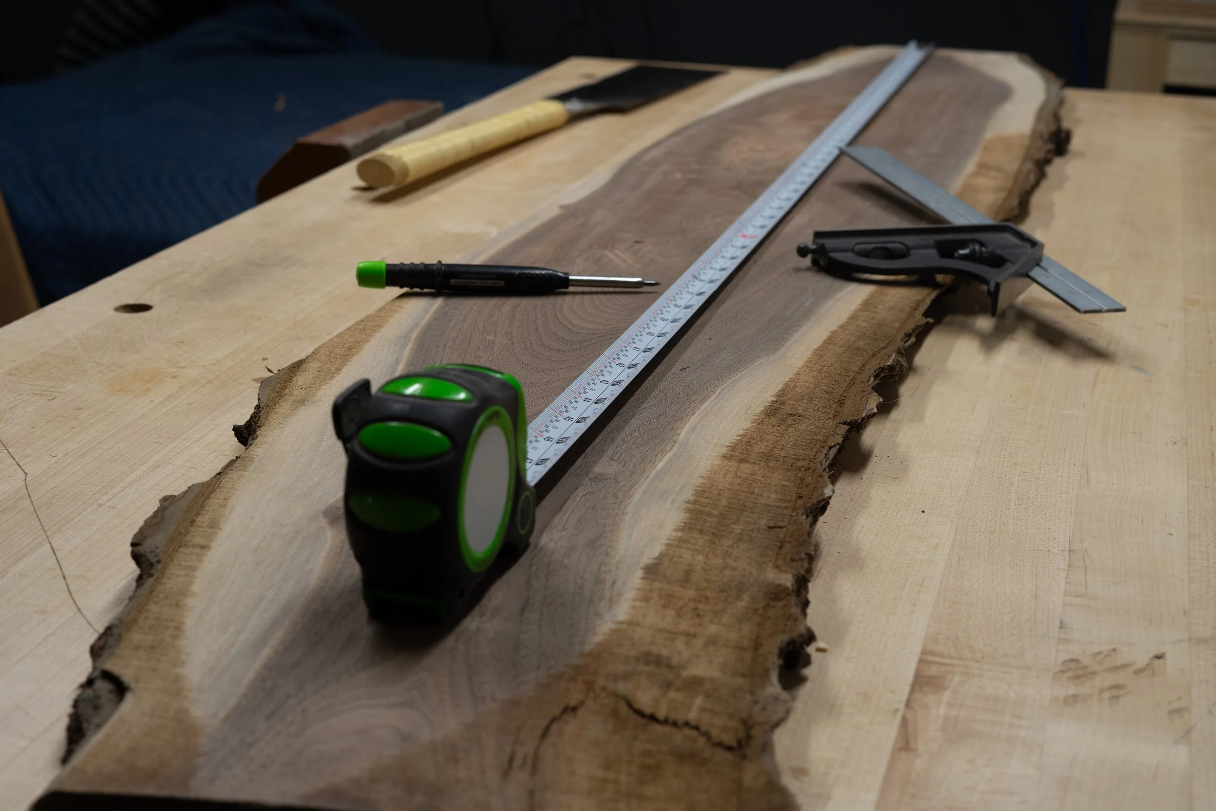 A woodworking workspace featuring a large piece of wood with tools including a measuring tape, a square, a ruler, a screwdriver, and a chisel placed on top.