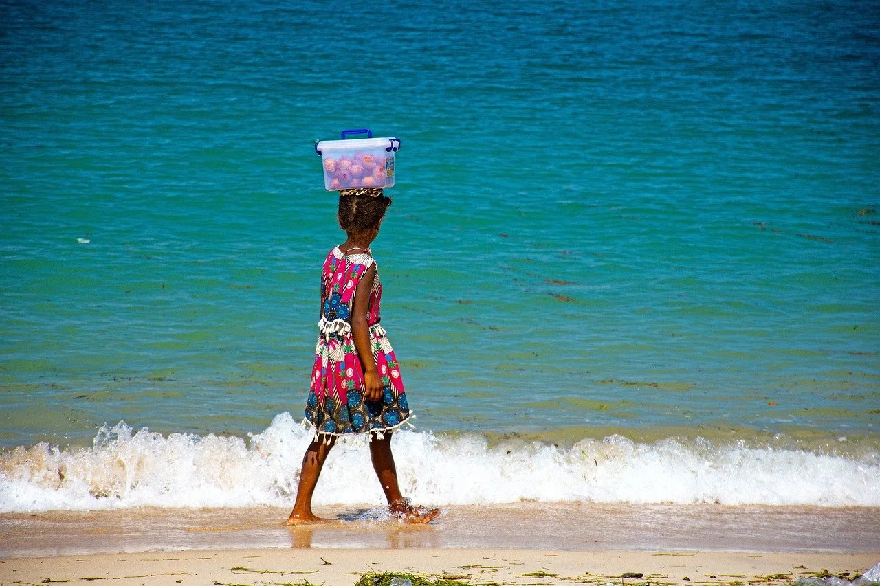 Child walking with a bin of apples on her head by the beach