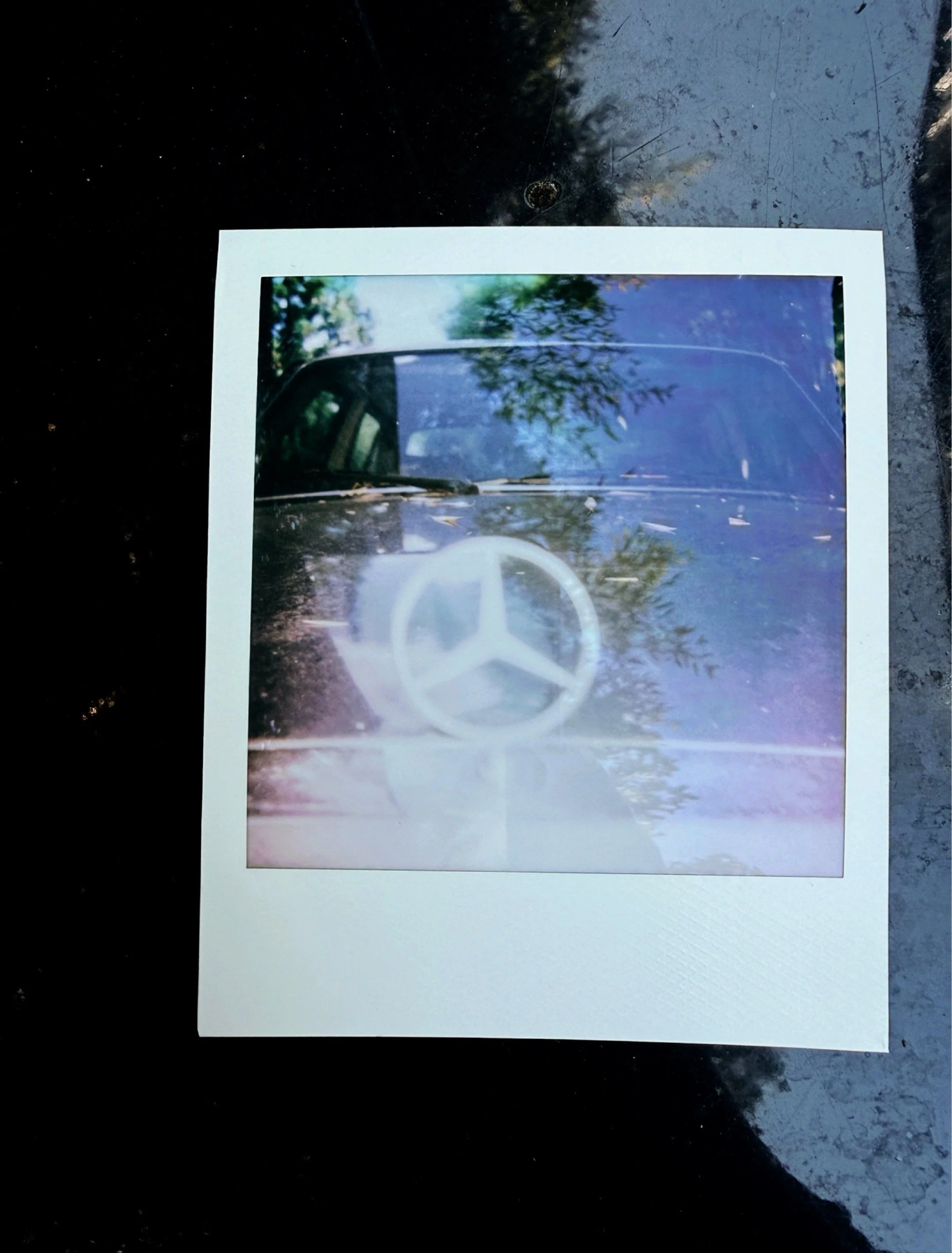 Polaroid photograph of a Mercedes hood ornament with purple and blue tones and tree reflections on the hood