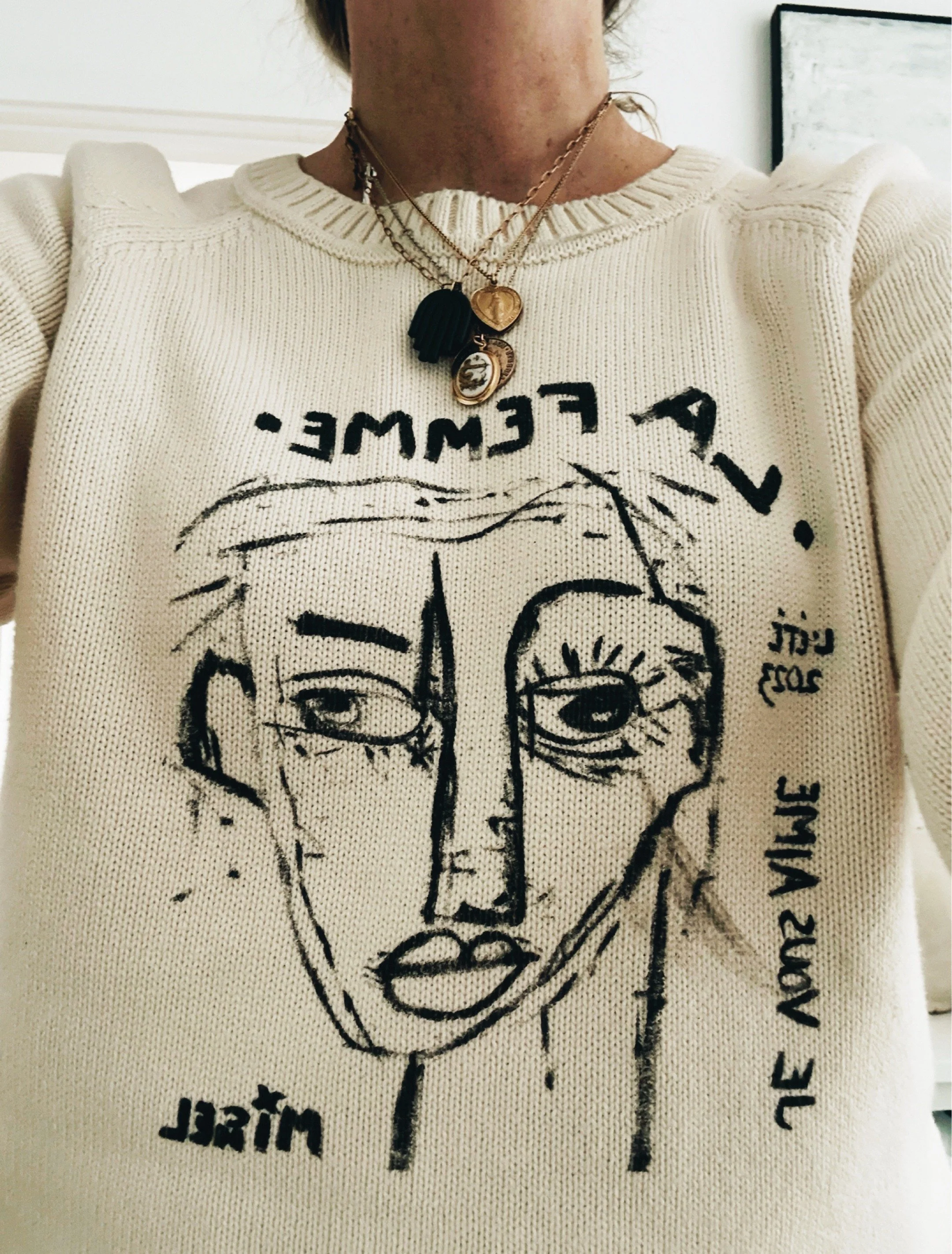 Cream sweater featuring an abstract black line drawing of a face by Michel Van Devender with layered necklaces.