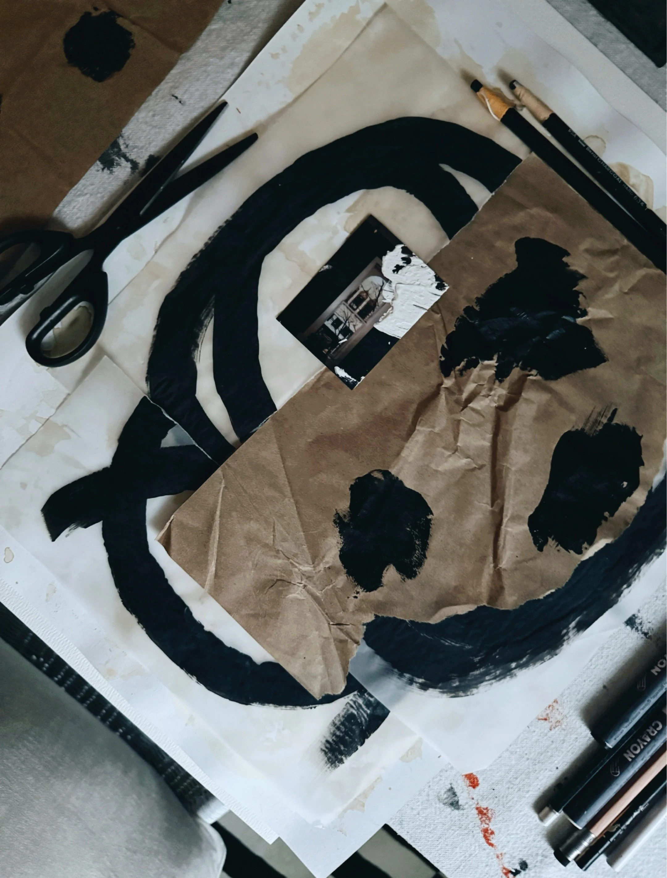 Abstract collage in progress with black brushstrokes on kraft paper scissors and charcoal pencils on studio table