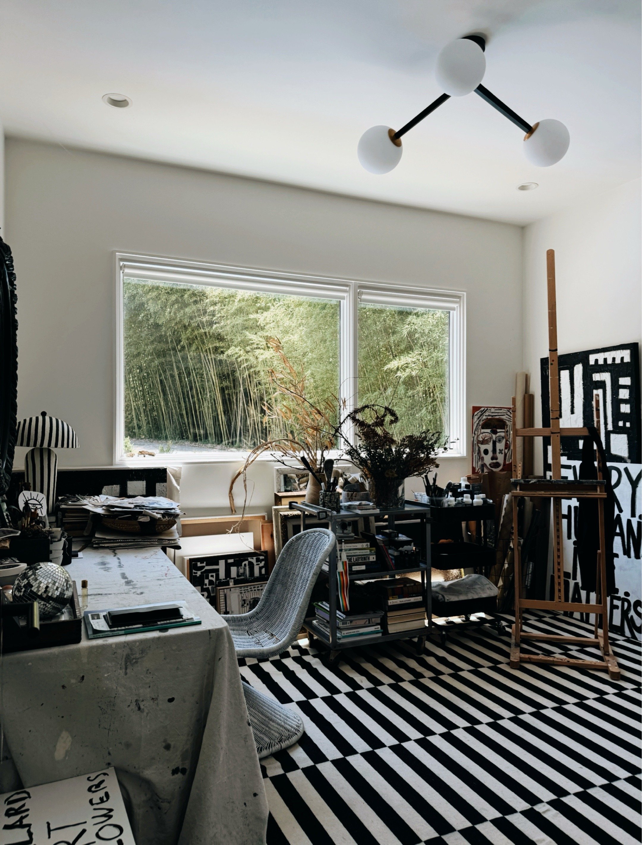 Art studio with black and white striped rug, framed artwork, potted bamboo, and natural light streaming through large windows.