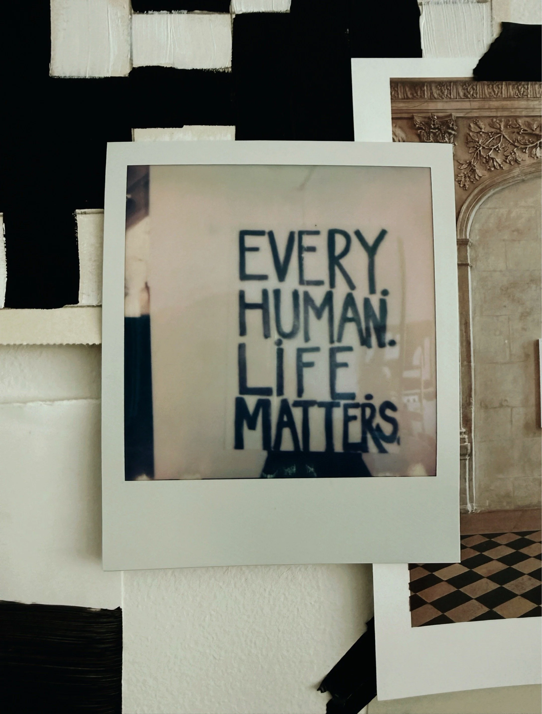 Polaroid photograph of a hand-lettered sign reading Every Human Life Matters, photographed against Michel Van Devender's black and white abstract paintings