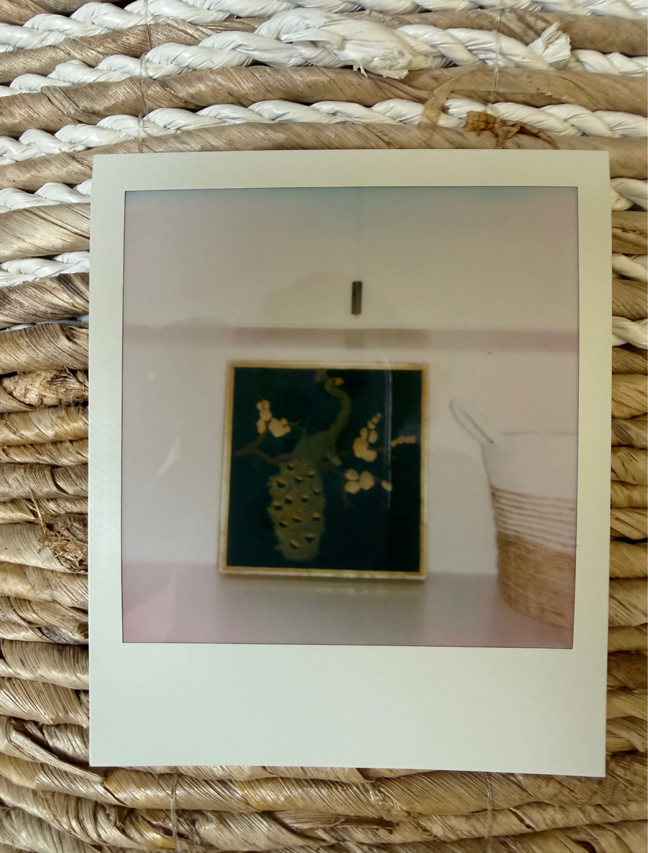 Polaroid photograph of a dark green framed painting on a white wall with a woven basket beside it