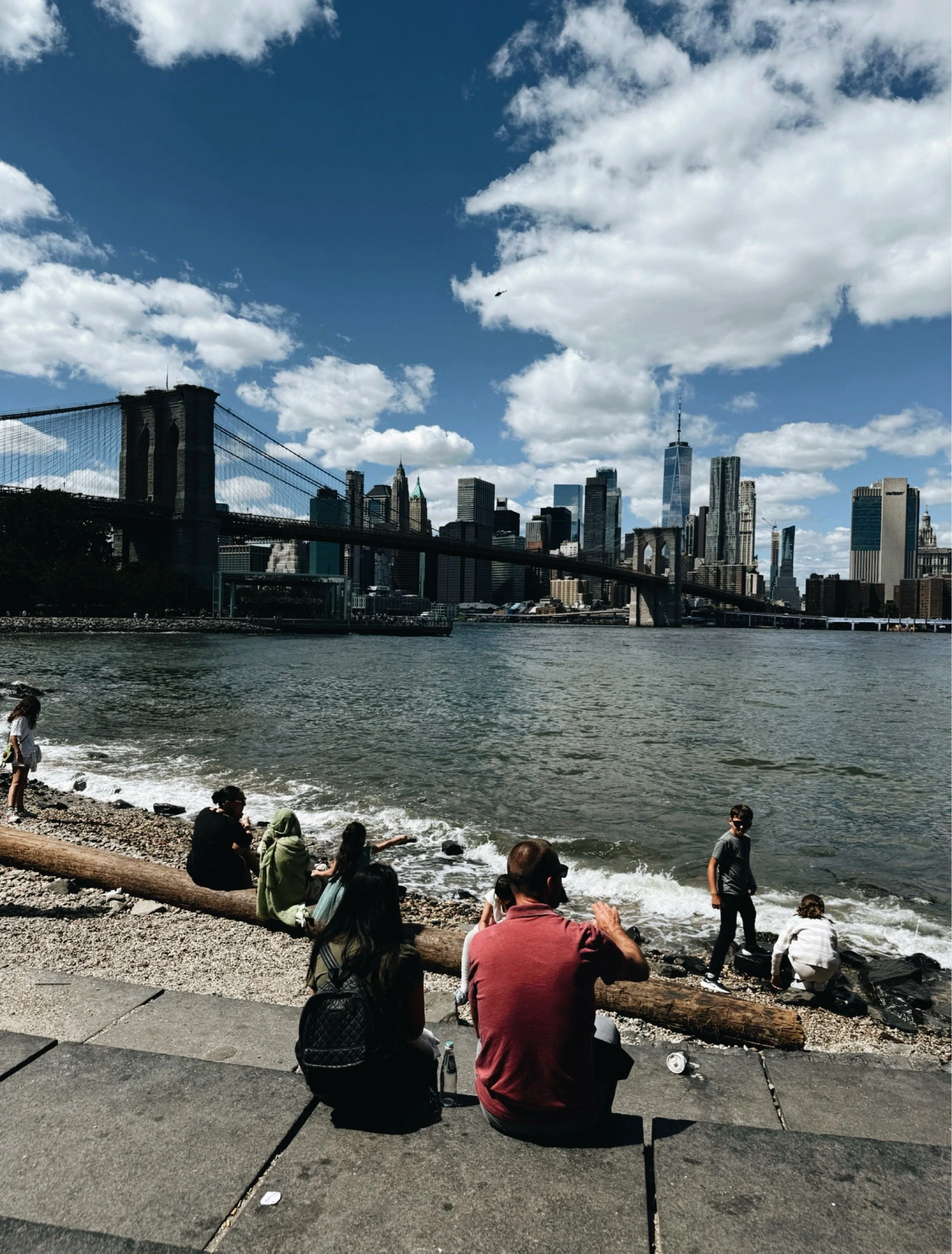 People sitting along the waterfront overlooking the Manhattan skyline and Brooklyn Bridge in New York City.