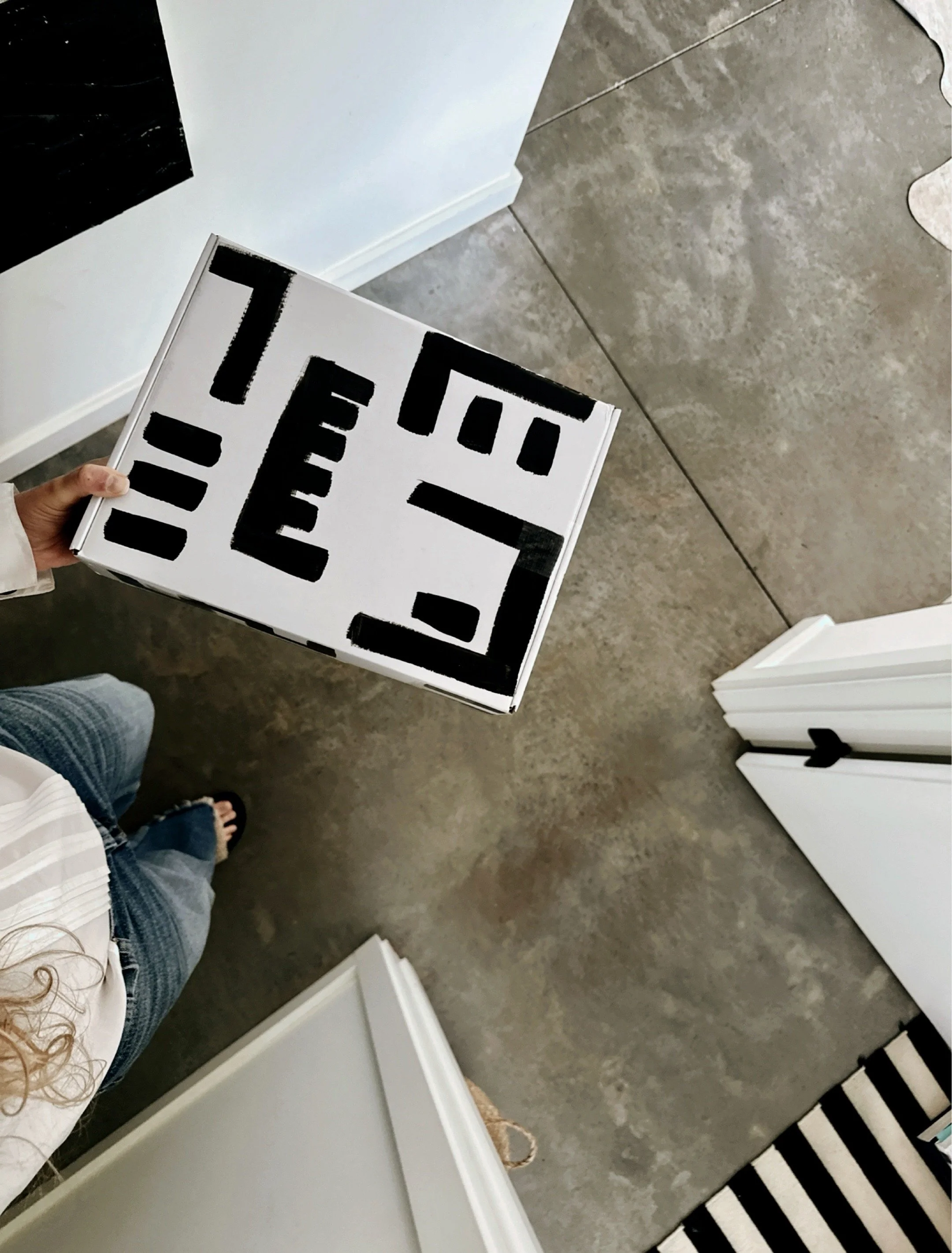 Hand holding a white box painted with bold black abstract geometric shapes. A shipment of goods and wearables by artist Michel Van Devender.