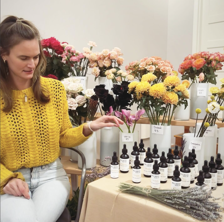 Woman in a yellow sweater sitting beside a table with small bottles and a variety of flower arrangements in vases.