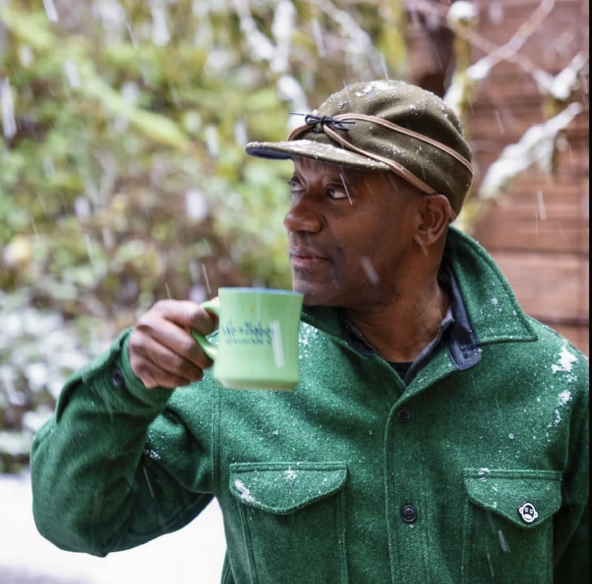 A man wearing a green jacket and a tan hat with glasses on the brim is holding a green mug and looking to the side outside in the rain.