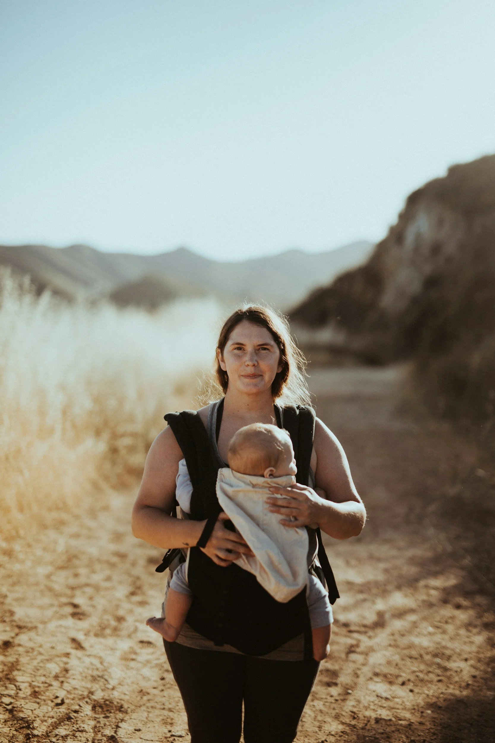 A mum hiking on a dirt trail in a mountainous area, carrying a baby in a front-facing baby carrier.