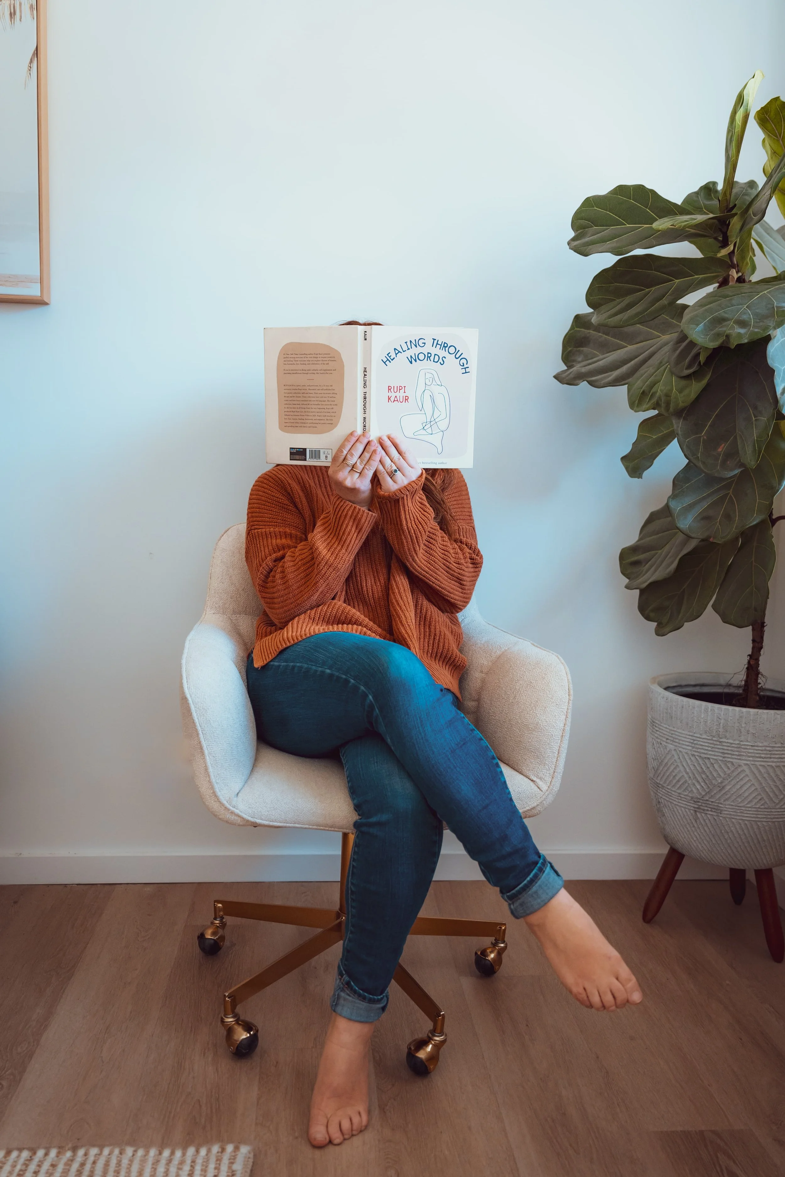 Person sitting on a cream-colored chair with a person underneath reading a book titled 'Healing through Words.' The person is wearing a rust-colored sweater and blue jeans, with one foot resting on the floor and the other crossed over the knee. There is a large potted plant on the right side of the image.