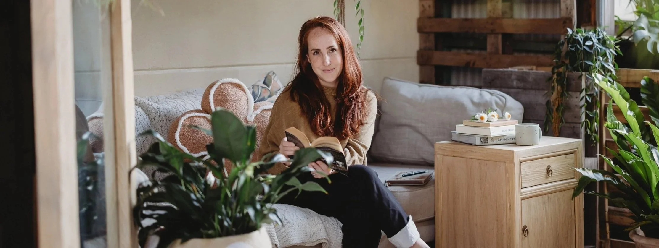 A counsellor for mums with red hair, wearing a beige sweater, sits on a light-colored couch in a cozy, plant-filled room, holding a book.