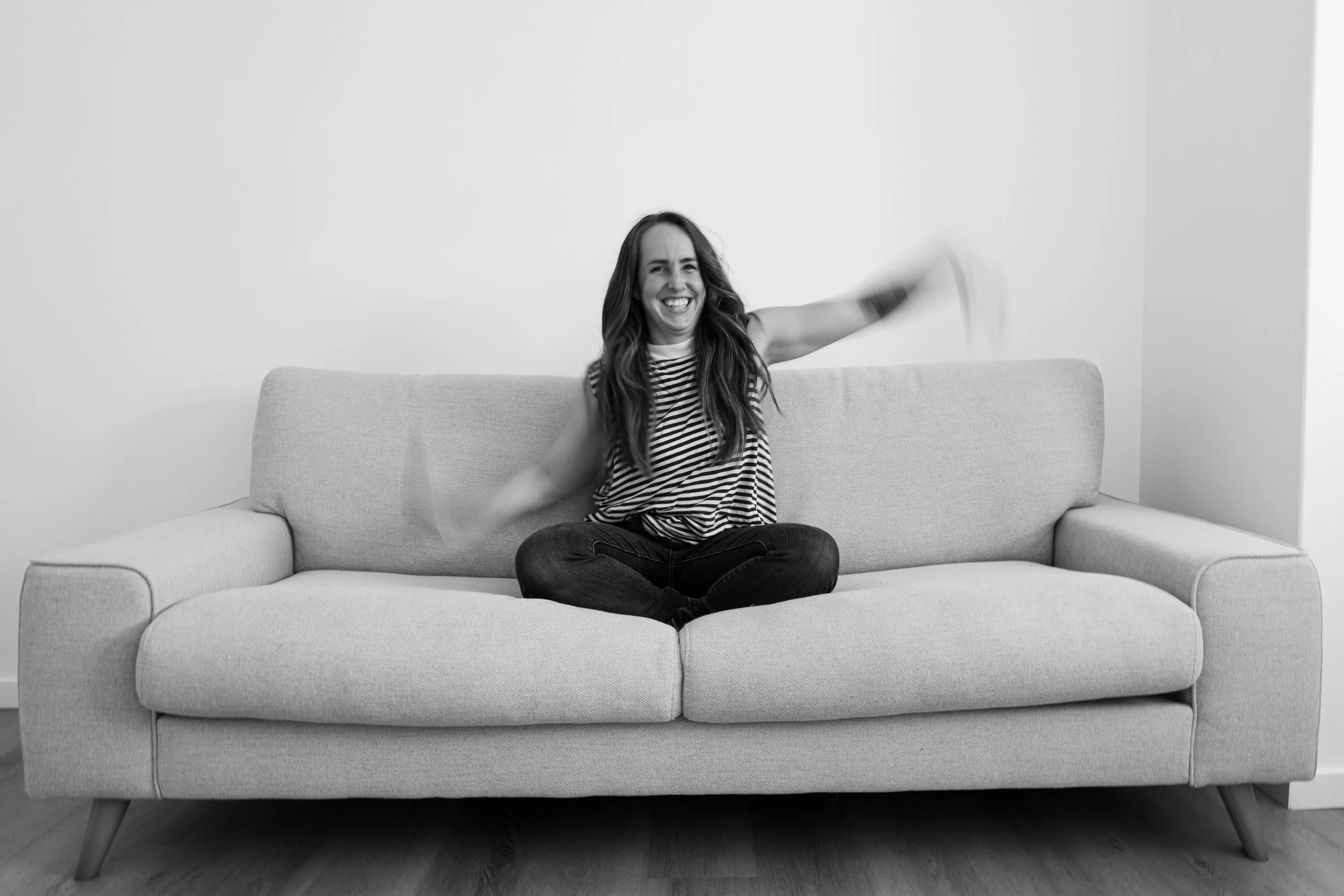 A woman sitting cross-legged on a light-colored sofa, smiling and waving with her right arm, wearing a striped shirt, in a minimalist room with plain white walls.