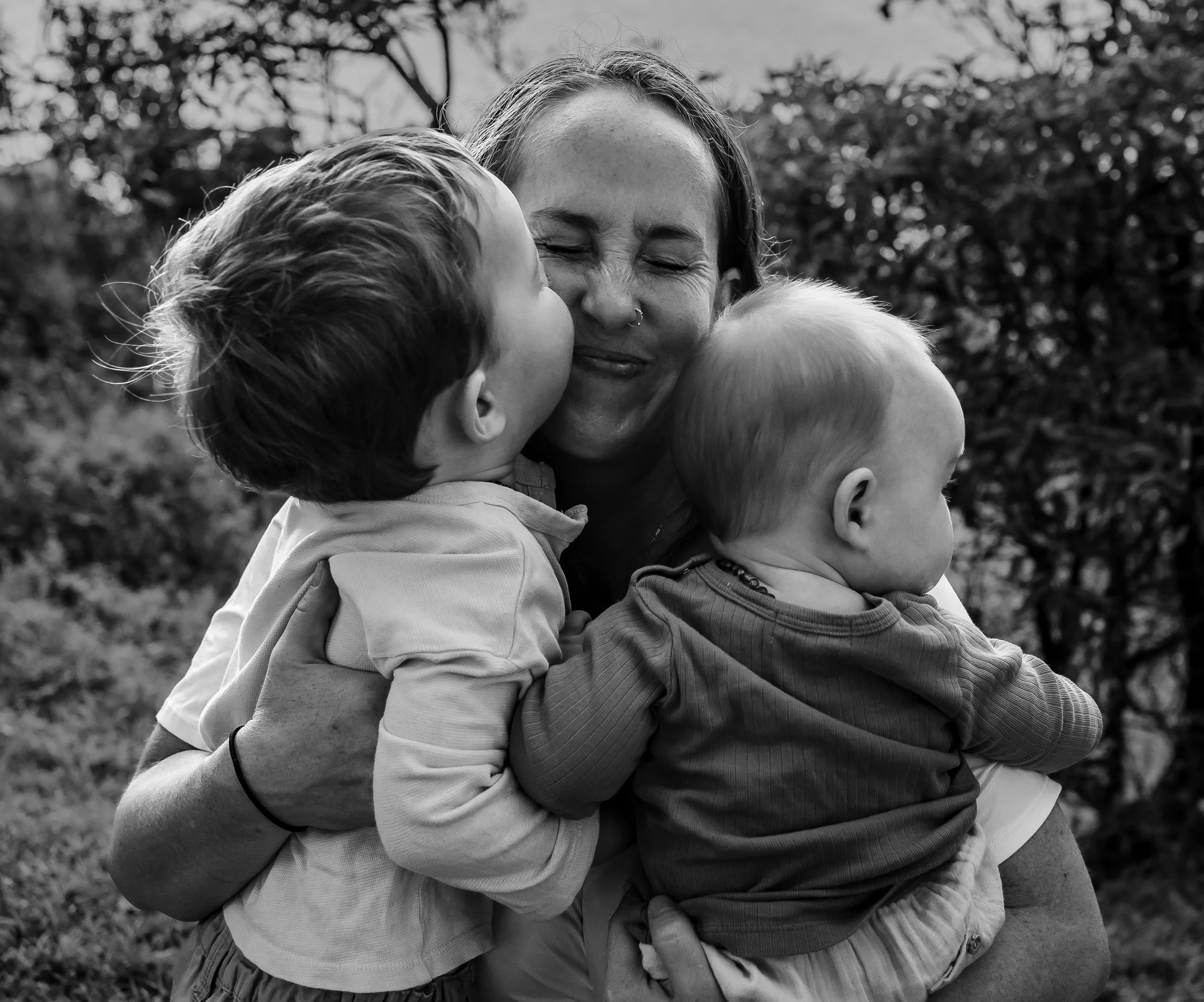 A mum hugging two young children outdoors, with one child giving her a kiss on the cheek. The scene is in black and white.