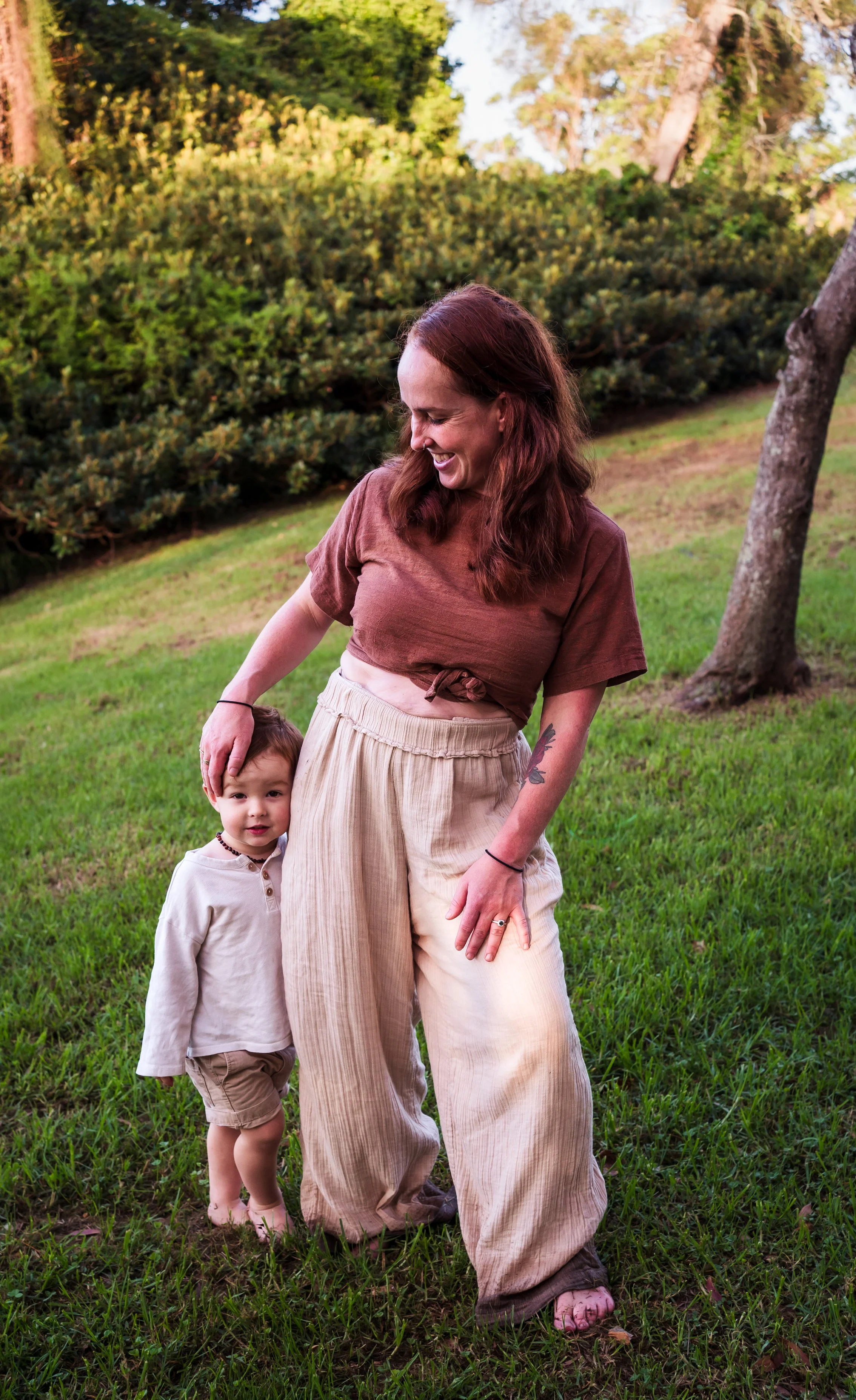 A mum and counsellor for mums with long red hair and a tattoo on her arm, smiling, standing outside on grass, with a young boy hugging her, both enjoying a sunny day in a park or backyard with trees and bushes in the background.
